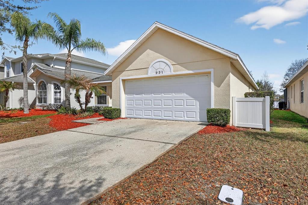 A single-story house exterior features a white garage door, tan stucco walls, and a driveway — 931 EMERALD GREEN COURT, KISSIMMEE, FL