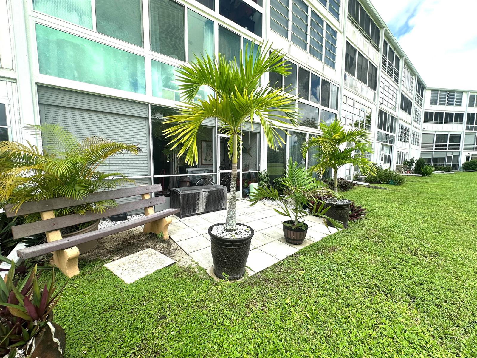 A patio with potted palm trees and a wooden bench sits outside a multi-story building — 1092 Newport S # 1092, Deerfield Beach FL 33442, DEERFIELD BEACH, FL
