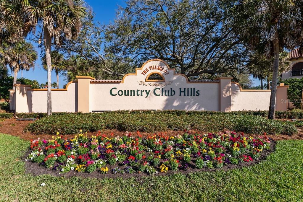 A beige monument sign for The Village of Country Club Hills sits behind flower beds — 1116 W BOONE COURT, LADY LAKE, FL