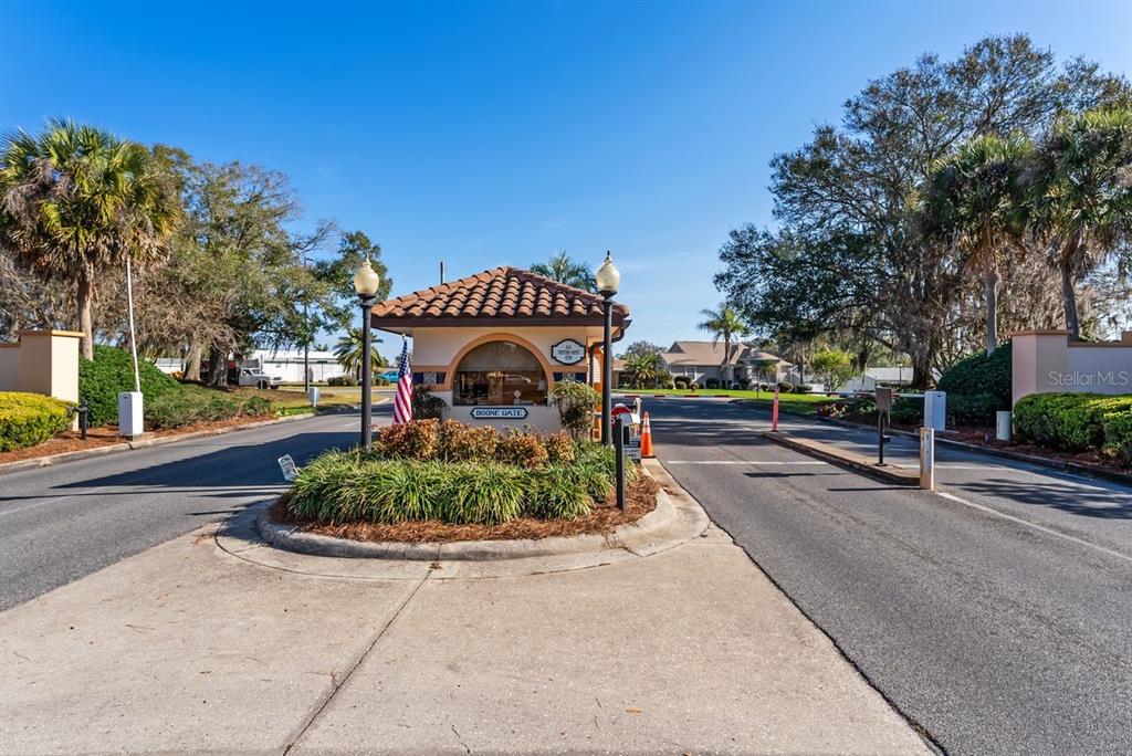 A gated community entrance features a guardhouse with a tile roof and landscaping — 1116 W BOONE COURT, LADY LAKE, FL
