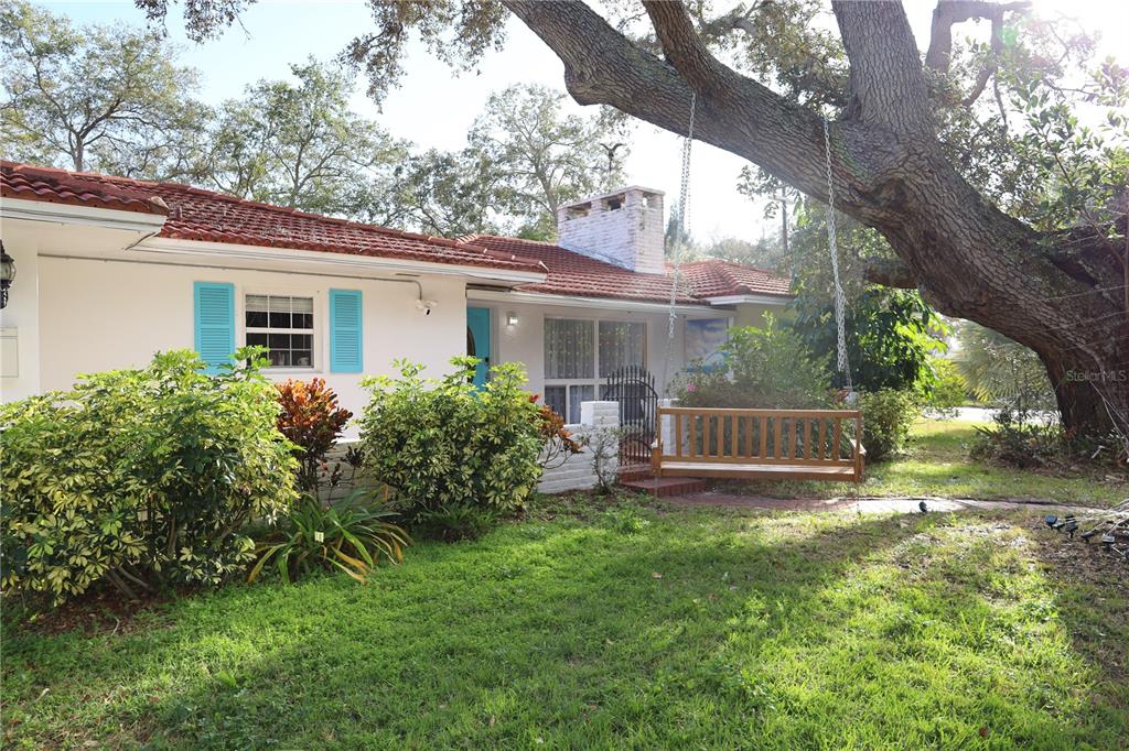 A white house with a red tile roof features a wooden swing under a tree — 7716 DARTMOUTH AVENUE N, ST PETERSBURG, FL