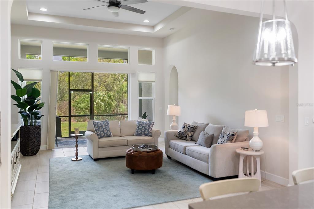 A living room features two beige sofas, a brown ottoman, and large windows overlooking trees — 2312 STARWOOD COURT, BRADENTON, FL