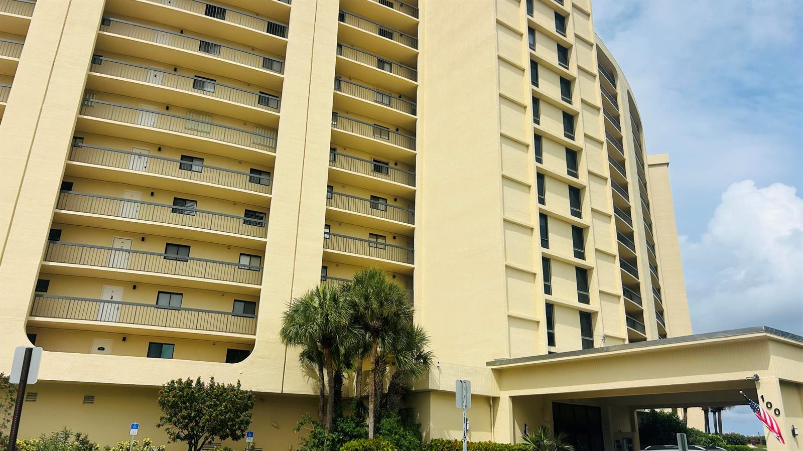 A low-angle exterior view of a tall, yellow multi-story apartment building with balconies — 100 Ocean Trail Way # 1002, Jupiter FL 33477, JUPITER, FL