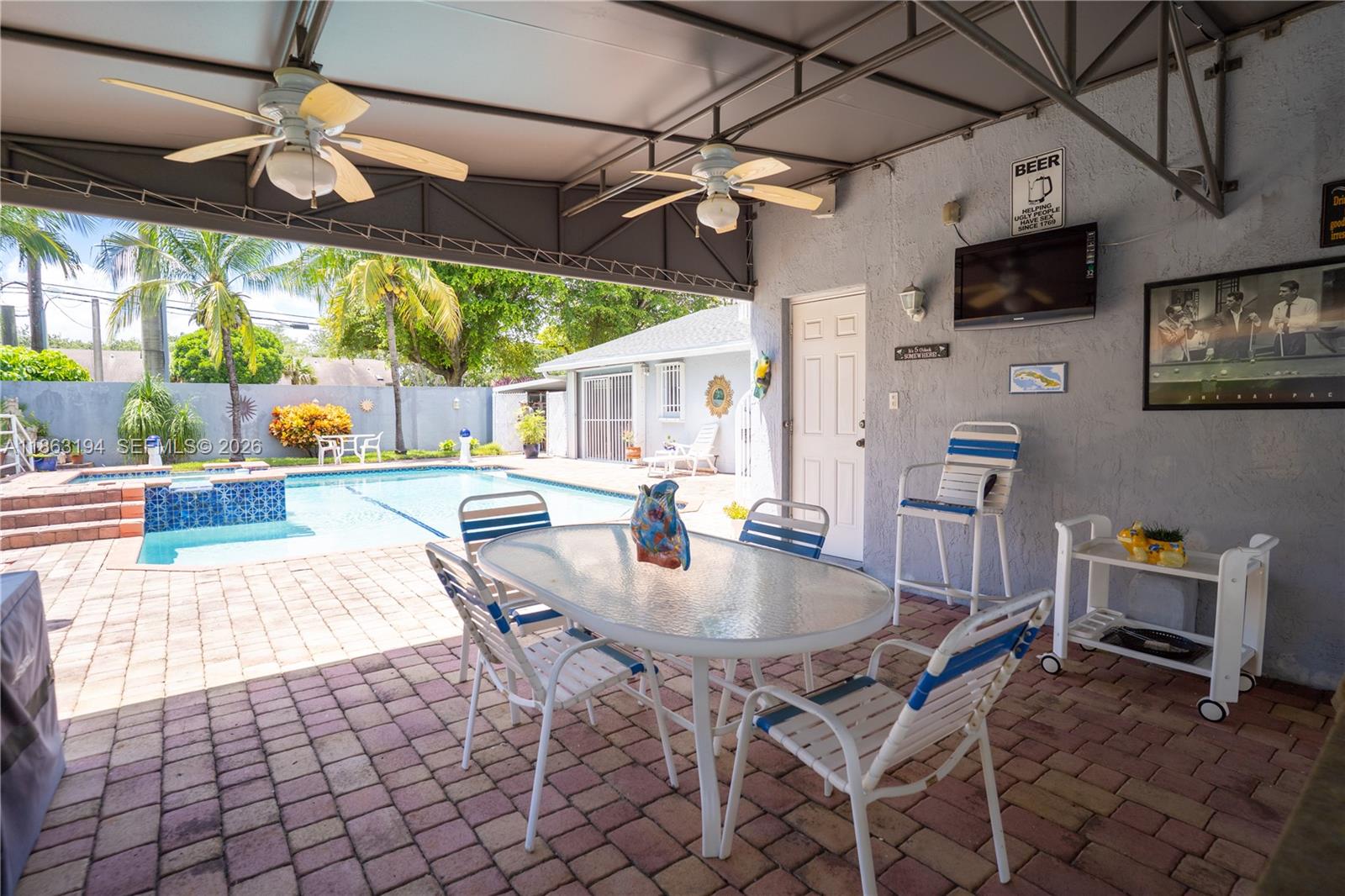 A covered patio features a dining set, pool view, and wall-mounted television on brick pavers — 11680 SW 92nd St, Miami FL 33176, MIAMI, FL