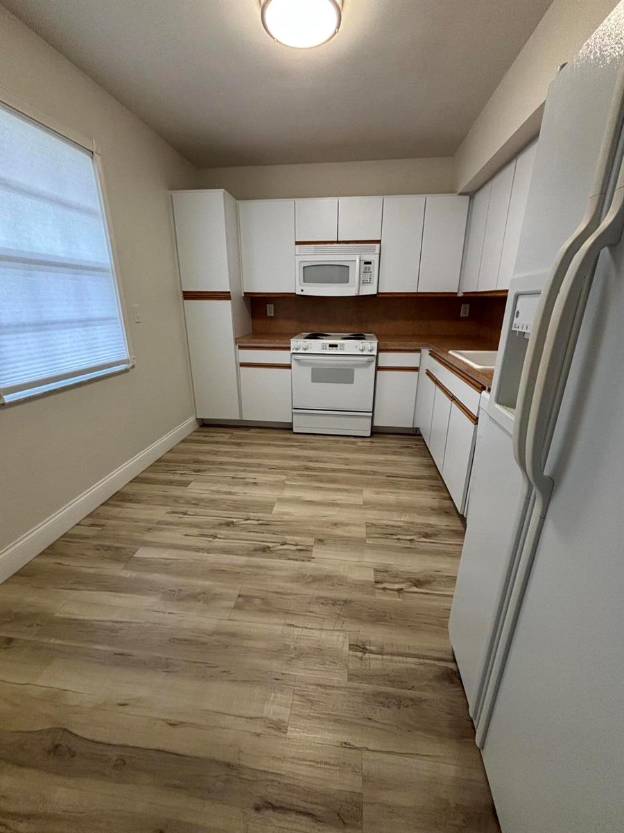 A kitchen features white cabinets, white appliances, wood-look flooring, and a window with blinds — 555 Oaks Lane # 309, Pompano Beach FL 33069, POMPANO BEACH, FL