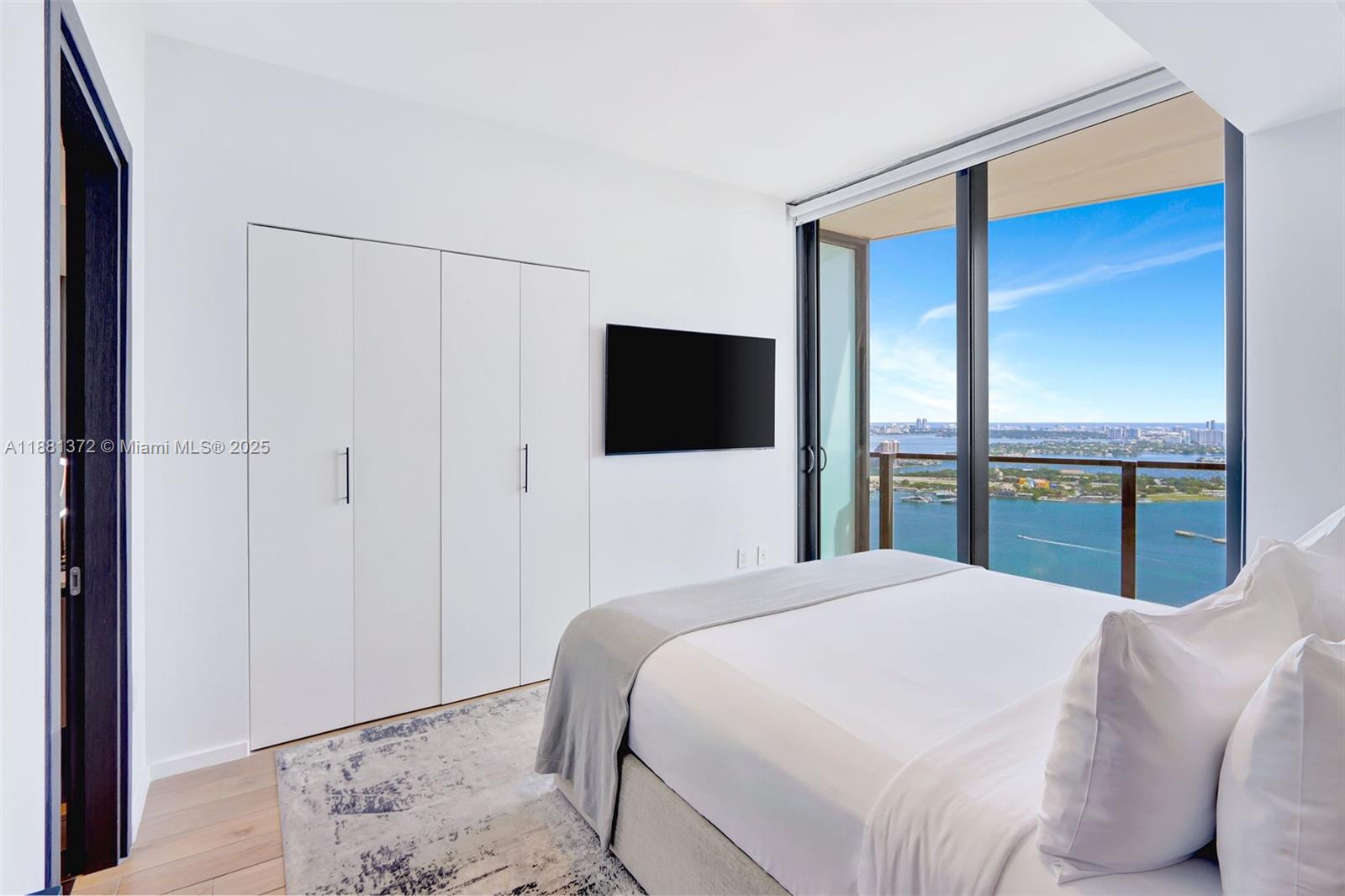 A bedroom features a bed, white closets, a wall-mounted television, and floor-to-ceiling windows overlooking water — 398 NE 5th St # 3810, Miami FL 33132, MIAMI, FL