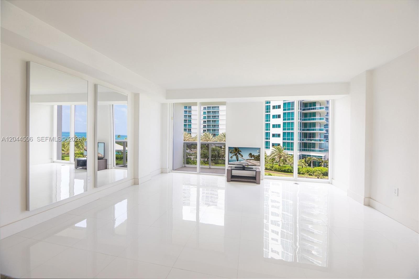 A bright, minimalist living room features white tile floors, large mirrors, and floor-to-ceiling windows — 10275 E Collins Ave # 502, Bal Harbour FL 33154, BAL HARBOUR, FL