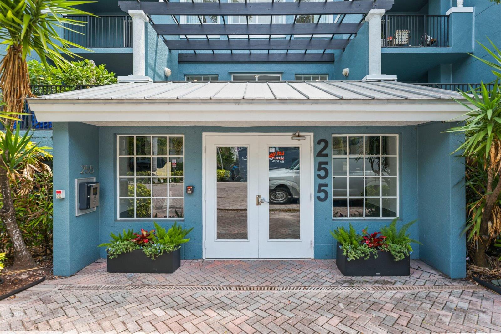 The exterior entrance of a blue building features a white door, windows, and a pergola — 255 NE 3rd Avenue Unit 407, Delray Beach FL 33444, DELRAY BEACH, FL