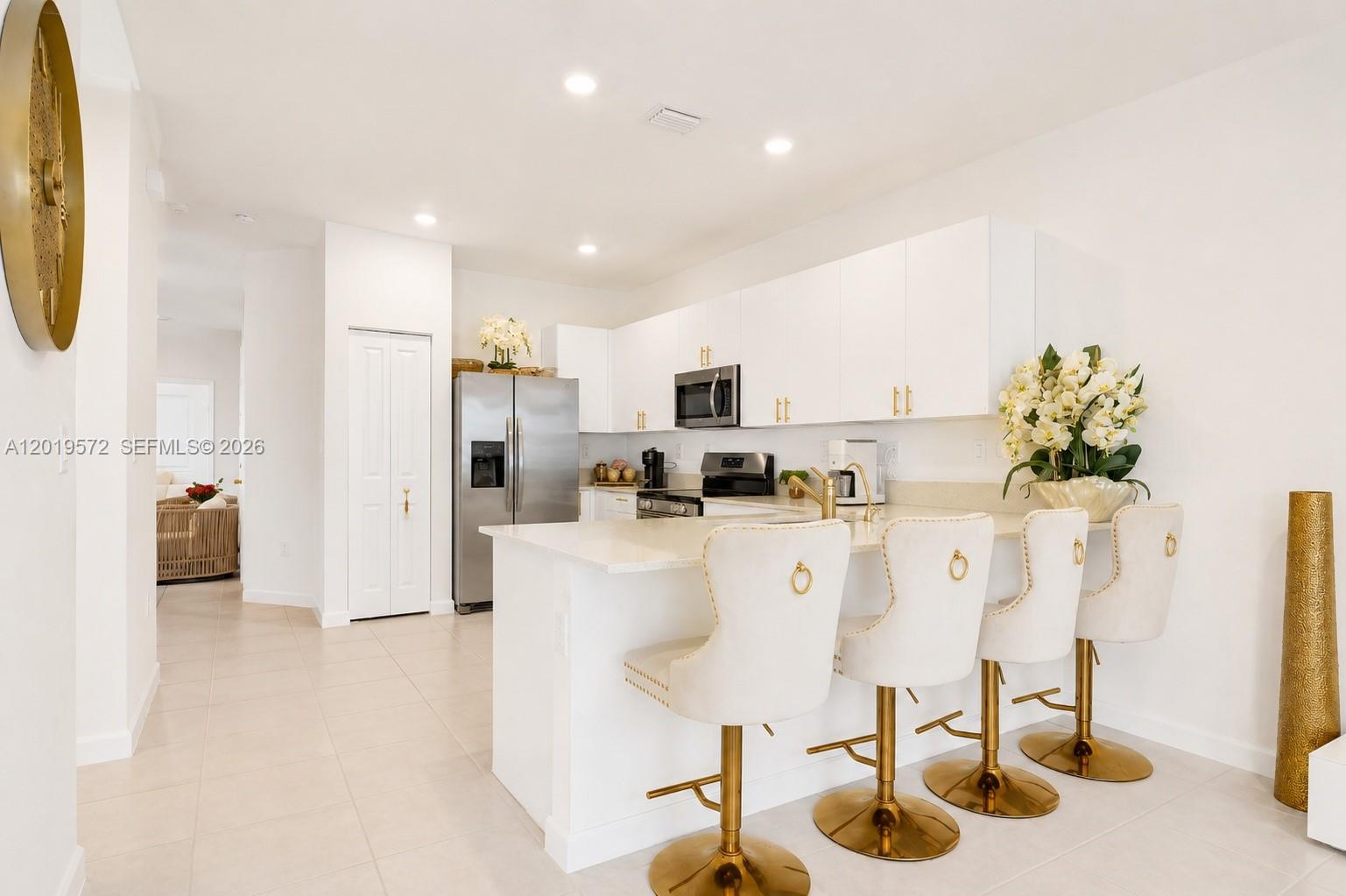 A modern white kitchen features a breakfast bar with four gold-accented stools — 12954 SW 231st St, Miami FL 33170, MIAMI, FL