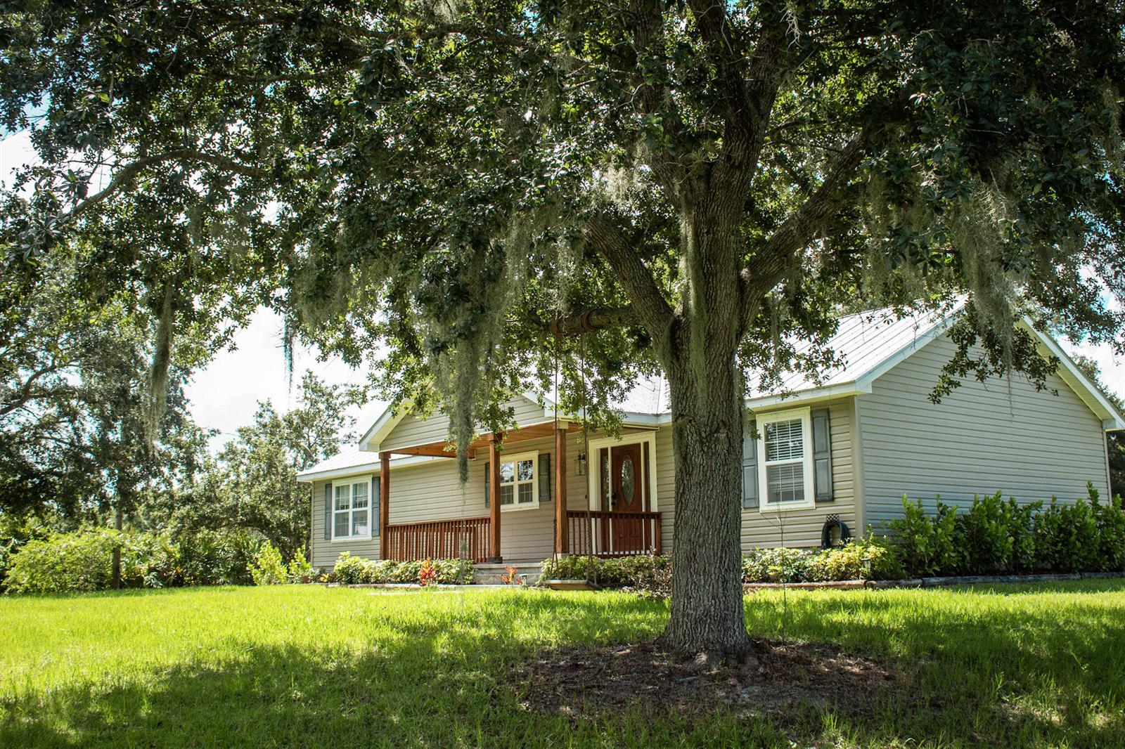 A single-story house with light siding and a front porch sits under a large tree — 24770 SW 95th Street, FL 34956, FL