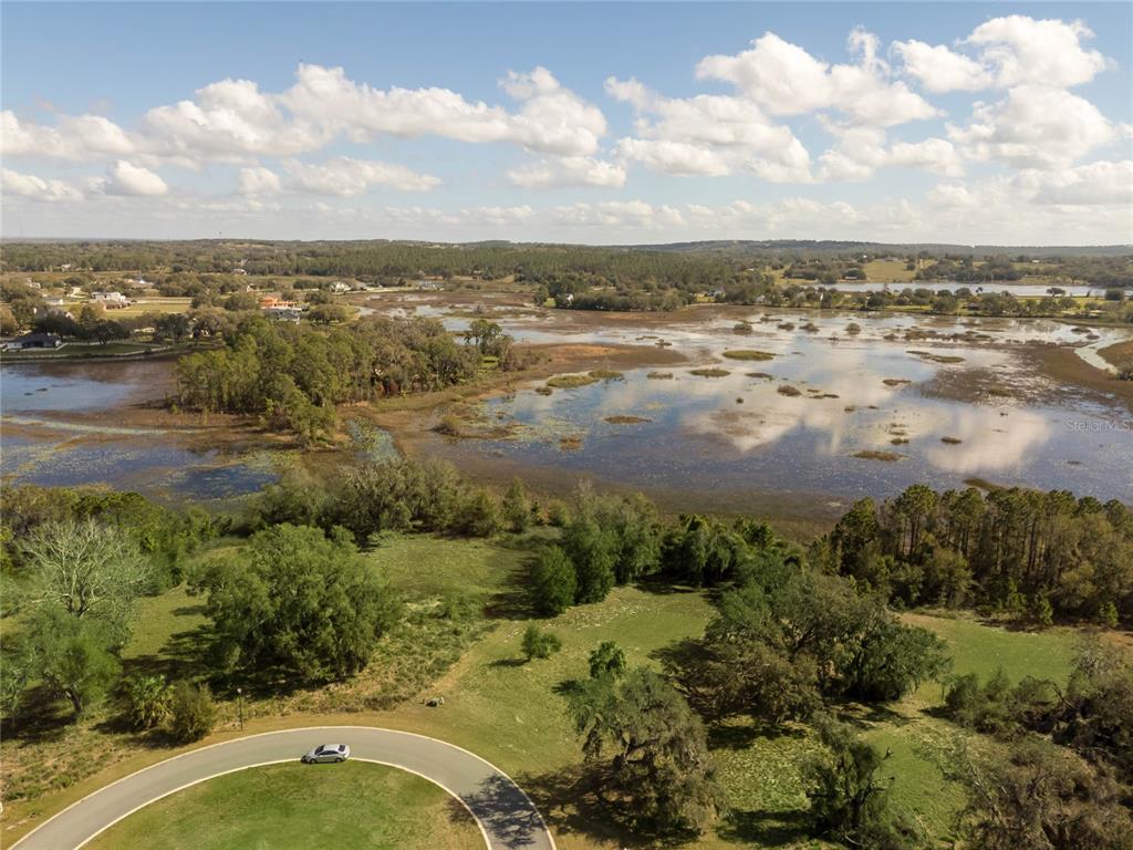 An aerial view shows a curved road bordering a grassy field and a large lake — 476 LONG AND WINDING RD, HOWEY IN THE HILLS, FL