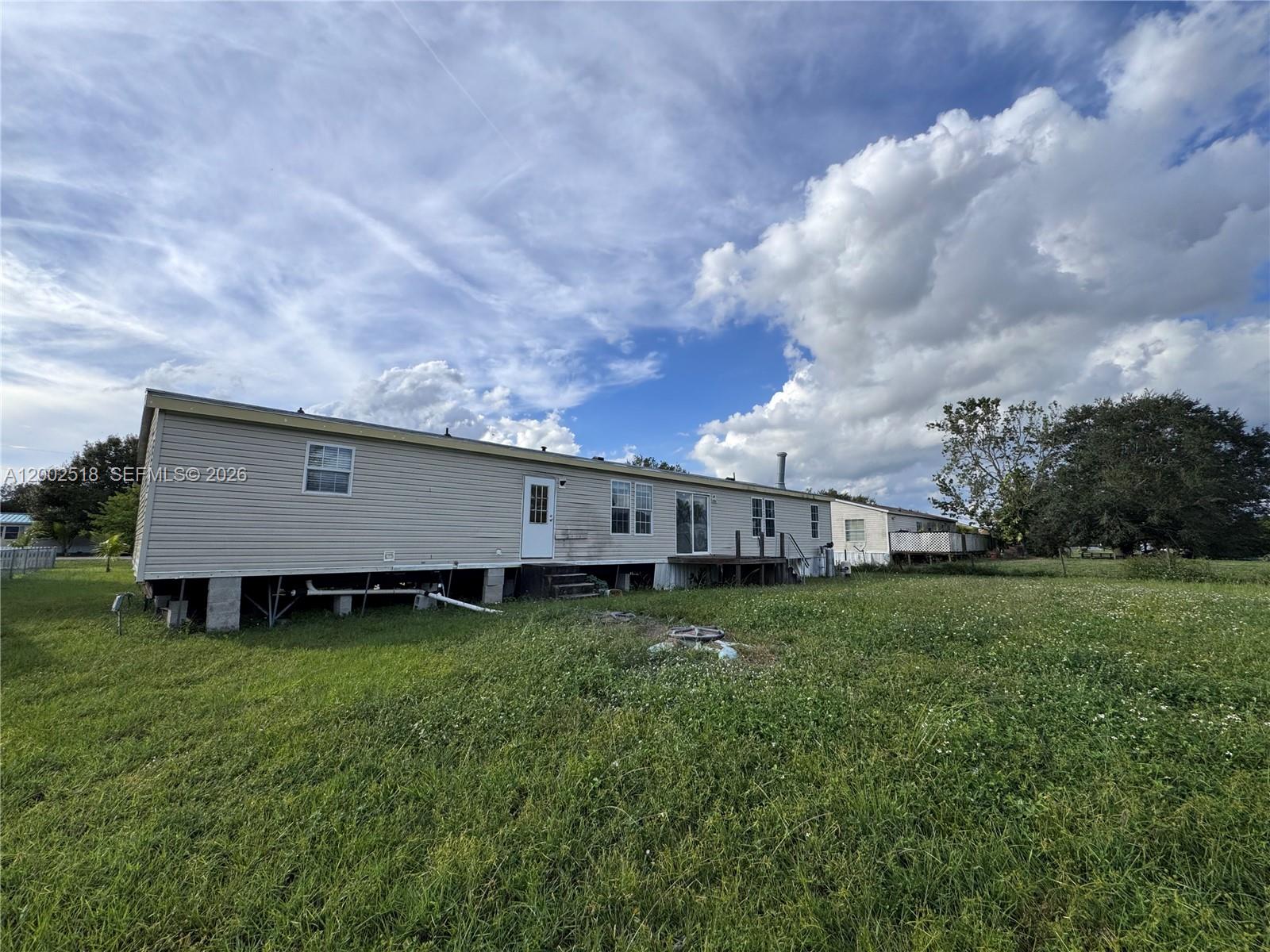 The exterior of a beige manufactured home sits on a grassy lot under a cloudy sky — 866 NE 104, Okeechobee FL 34972, OKEECHOBEE, FL