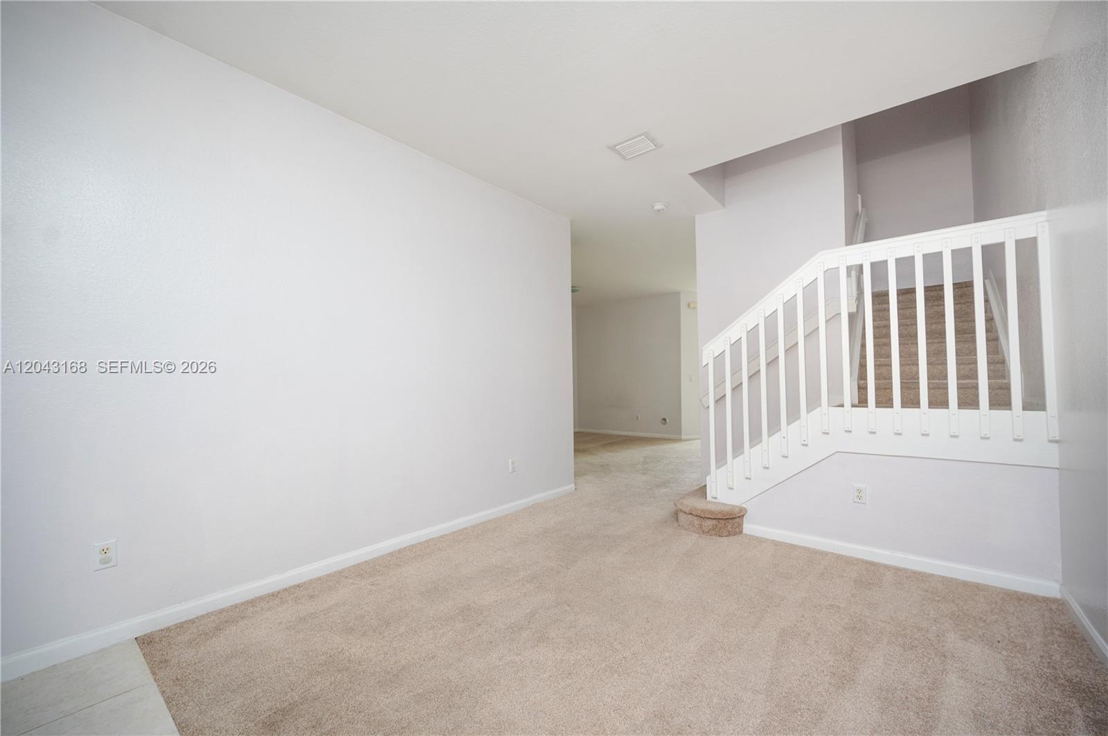 An empty room features beige carpeting, white walls, and a white staircase with a railing — 445 NE 21st Ave, Homestead FL 33033, HOMESTEAD, FL