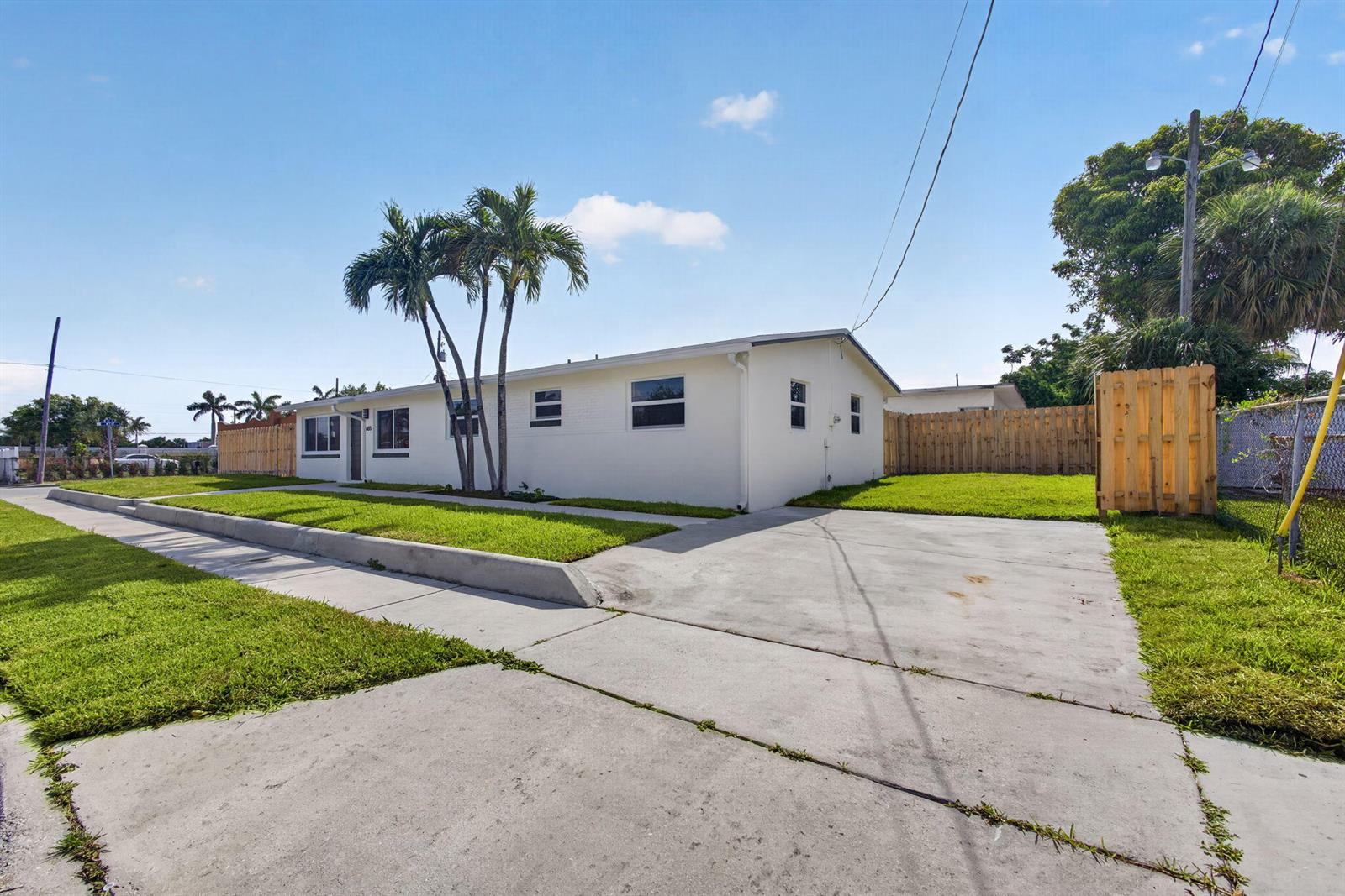 A single-story white house with a concrete driveway, palm trees, and a wooden fence — 665 W 7th Street, Riviera Beach FL 33404, RIVIERA BEACH, FL