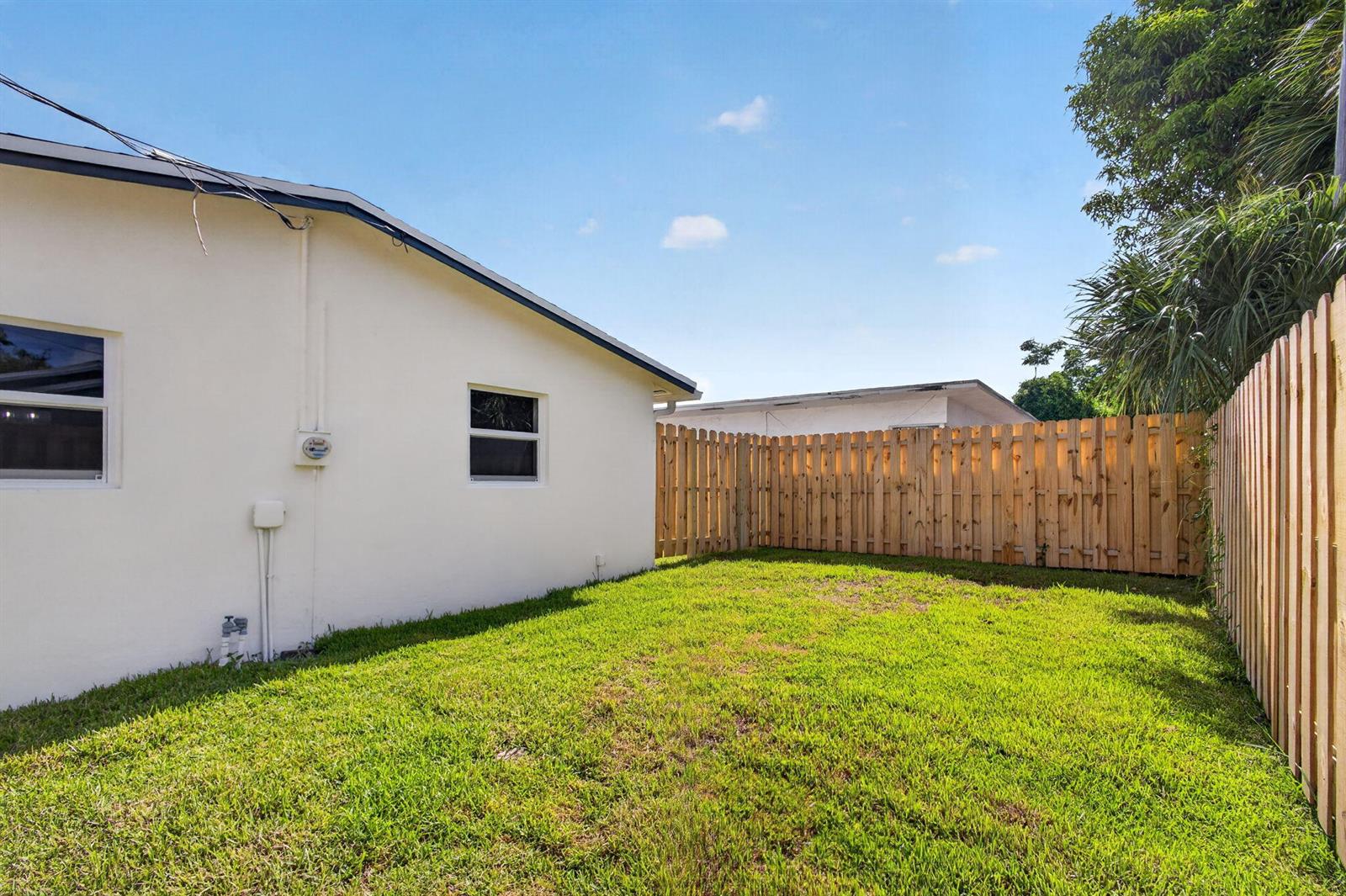 A side yard with a white house wall, green grass, and a wooden privacy fence — 665 W 7th Street, Riviera Beach FL 33404, RIVIERA BEACH, FL
