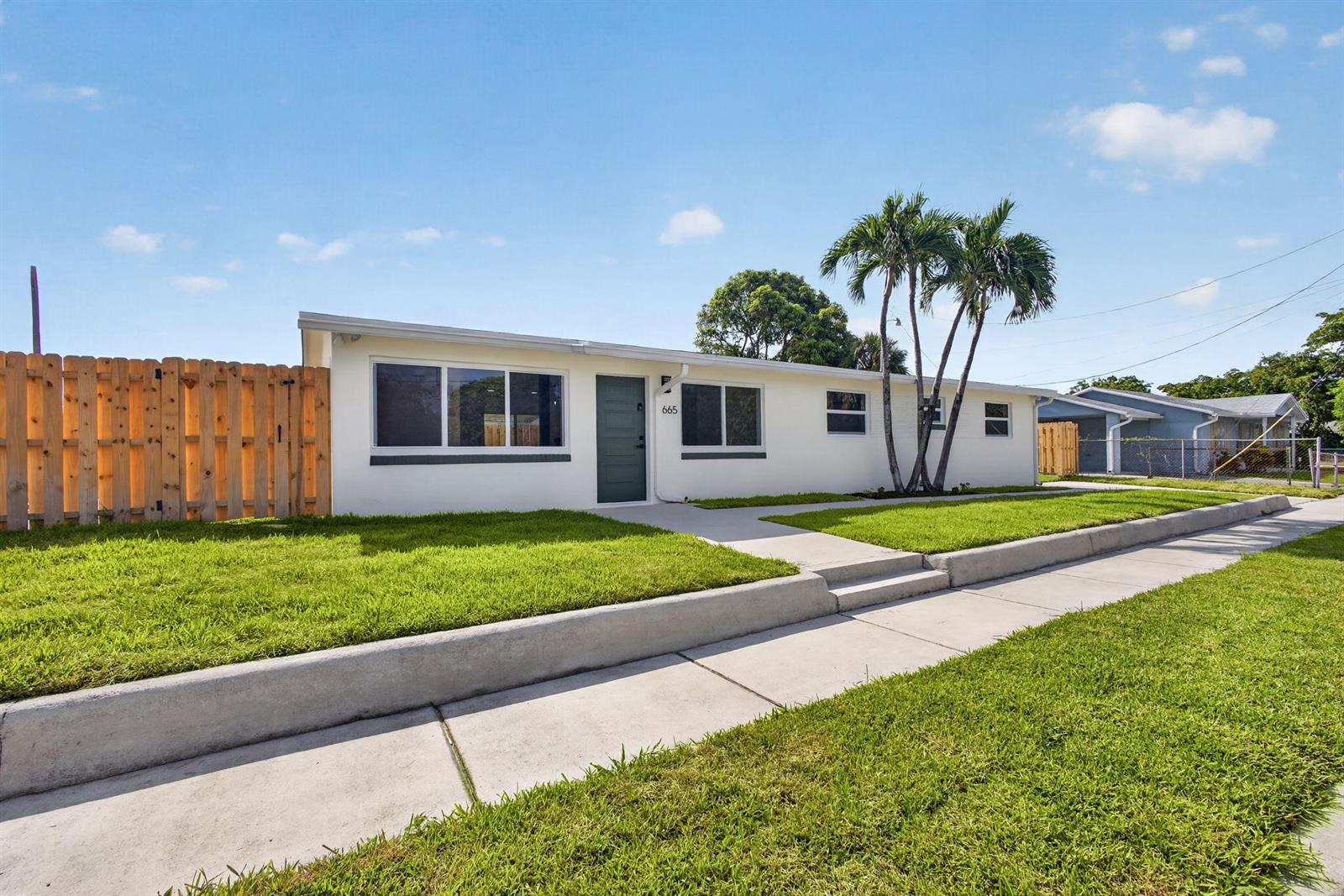 A single-story white house with a green door, palm trees, and a wooden fence — 665 W 7th Street, Riviera Beach FL 33404, RIVIERA BEACH, FL