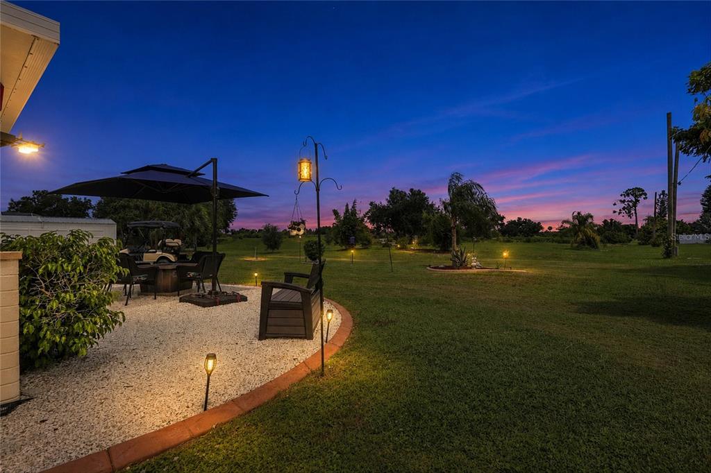 A patio with a table, umbrella, and landscape lighting overlooks a grassy yard at sunset — 22580 BOLANOS COURT, PORT CHARLOTTE, FL