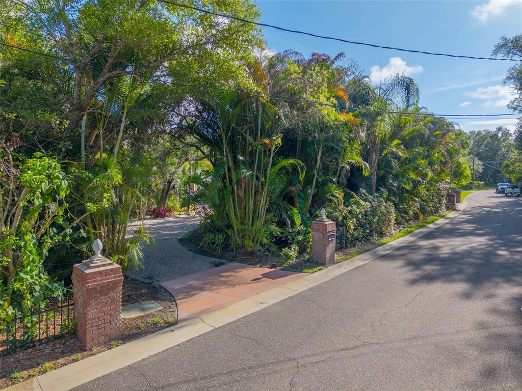 A brick-pillared driveway entrance leads into a lush, tree-filled property from a paved road — 1821 OAK CREEK DRIVE, DUNEDIN, FL