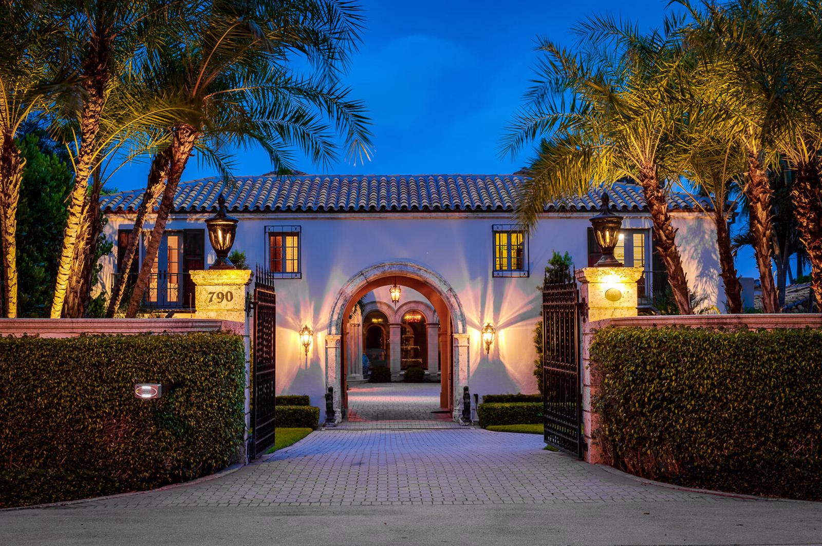 A gated Mediterranean-style home with a stone archway, paved driveway, and illuminated palm trees — 790 S County Road, Palm Beach FL 33480, PALM BEACH, FL
