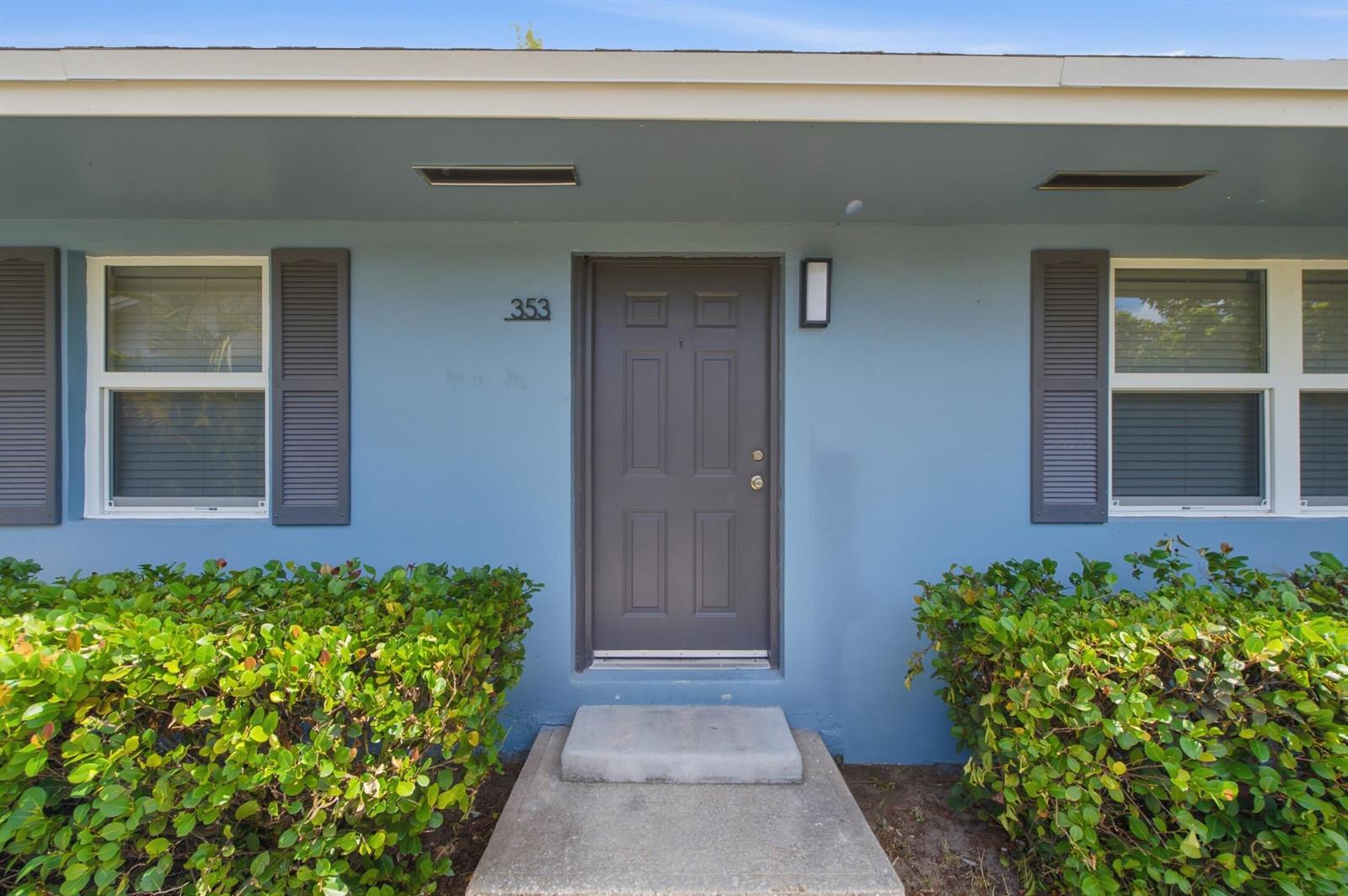 A blue exterior wall features a dark gray front door, two windows with shutters, and landscaping — 353 Wickline Boulevard, Lantana FL 33462, LANTANA, FL