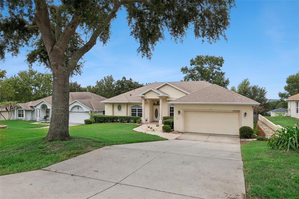 A single-story house with a beige stucco exterior, a two-car garage, and a large tree — 10308 CALLE DE FLORES DRIVE, CLERMONT, FL