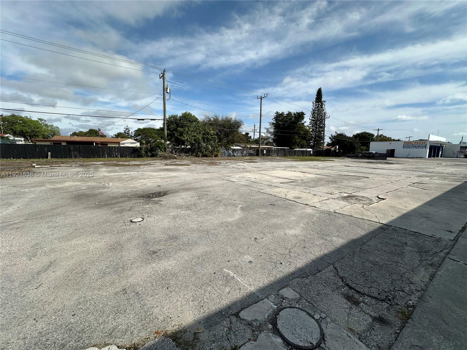 A large, vacant concrete lot is surrounded by a fence, trees, and nearby buildings — 6501 Pembroke Rd, Hollywood FL 33023, HOLLYWOOD, FL