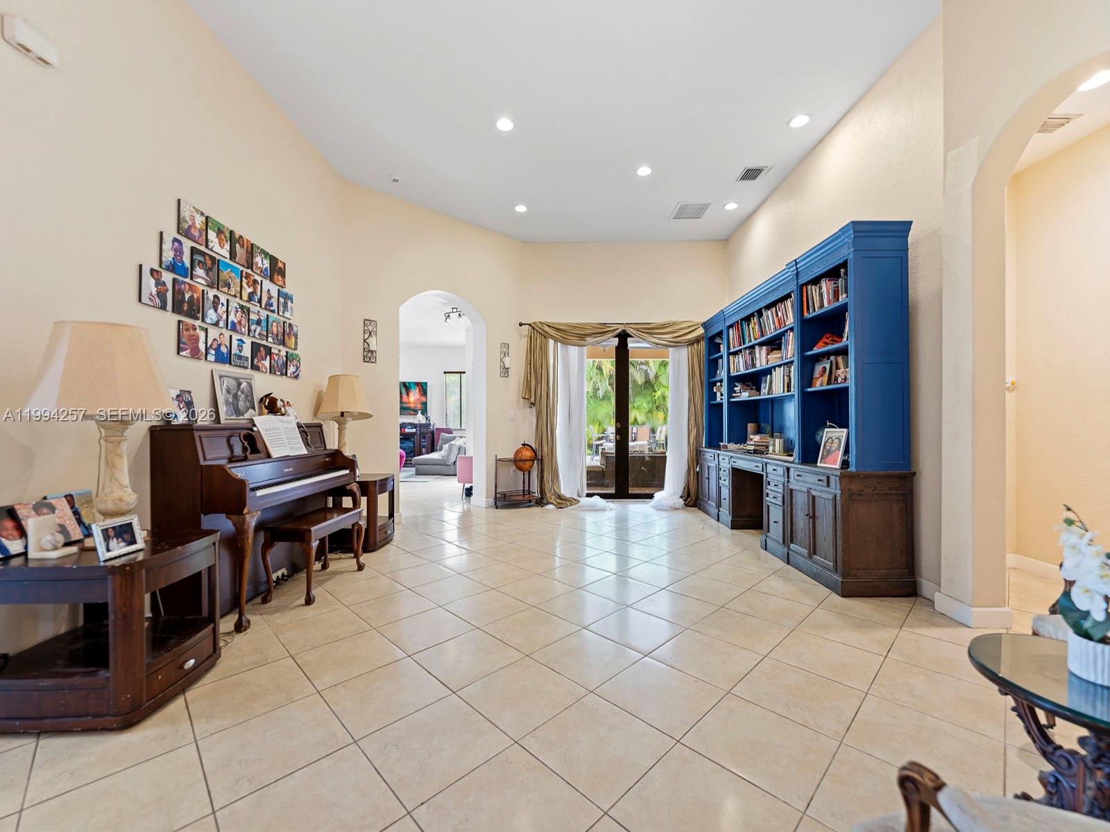 A spacious room features tile floors, a piano, a blue bookshelf, and an arched doorway — 19385 SW 79th Ct, Cutler Bay FL 33157, CUTLER BAY, FL
