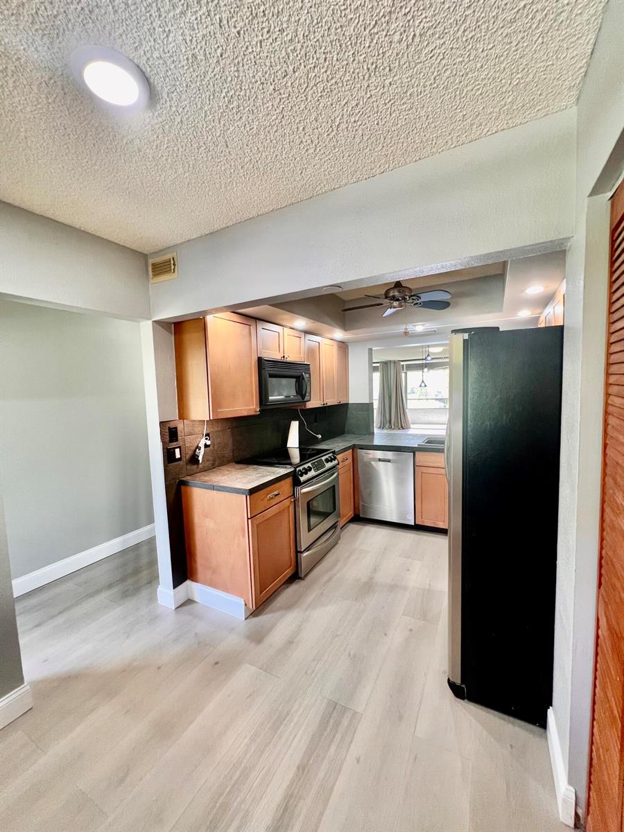 A kitchen features wood cabinets, stainless steel appliances, dark countertops, and light-colored wood-look flooring — 1850 Homewood Boulevard, Delray Beach FL 33445, DELRAY BEACH, FL