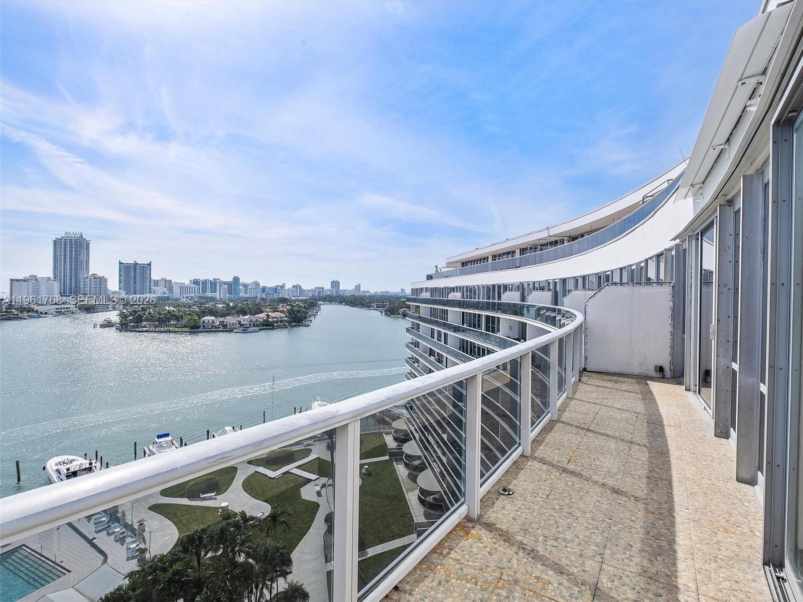 A curved balcony with glass railings overlooks a waterway, a park, and a city skyline — 900 Bay Dr # 1012, Miami Beach FL 33141, MIAMI BEACH, FL