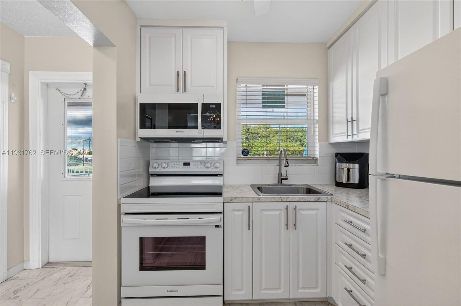 A small kitchen features white cabinets, white appliances, a stainless steel sink, and tile backsplash — 2650 W Golf Blvd # 259, Pompano Beach FL 33064, POMPANO BEACH, FL