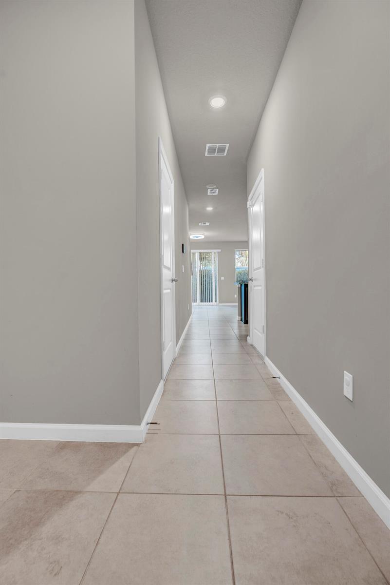 A long hallway with light gray walls, tile flooring, and white doors leads to a window — 5659 Enclave Drive, Riviera Beach FL 33407, RIVIERA BEACH, FL
