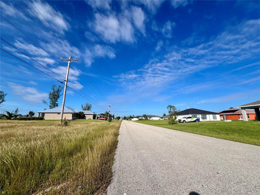 A paved road runs between a grassy vacant lot and residential houses under a blue sky — 1408 NE 37TH STREET, CAPE CORAL, FL