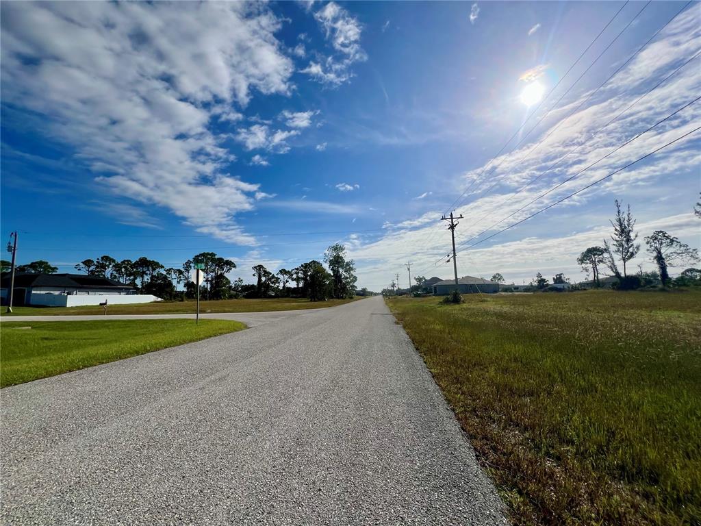 A paved road stretches through a grassy, undeveloped residential area under a sunny blue sky — 1408 NE 37TH STREET, CAPE CORAL, FL