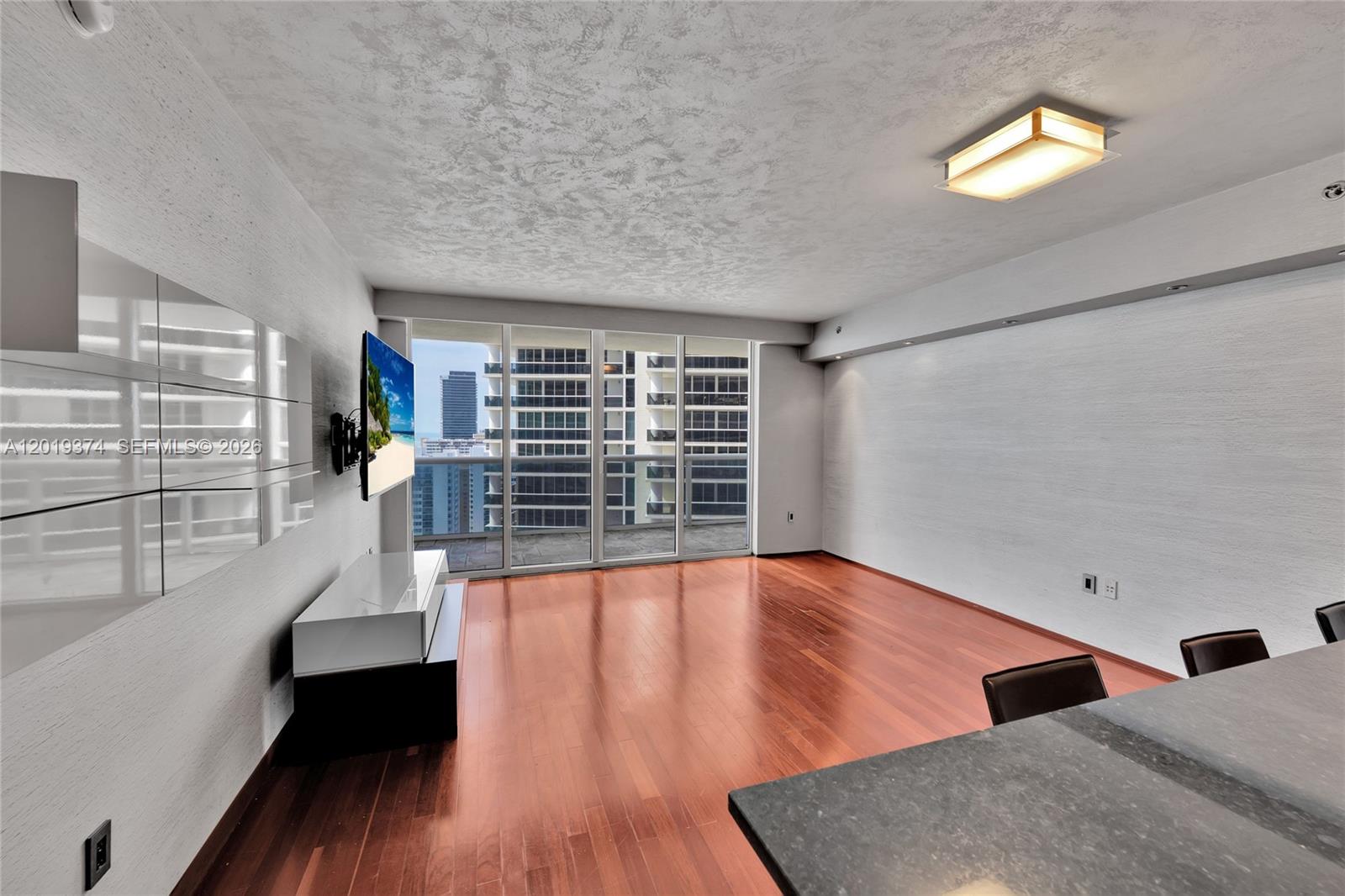 A living room with hardwood floors, a wall-mounted television, and floor-to-ceiling windows overlooking buildings — 1830 S Ocean Dr # 2705, Hallandale Beach FL 33009, HALLANDALE BEACH, FL