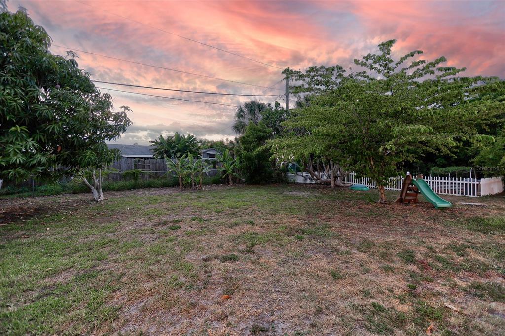 A grassy backyard features a small slide, mature trees, and a white picket fence — 258 READING STREET NW, PORT CHARLOTTE, FL