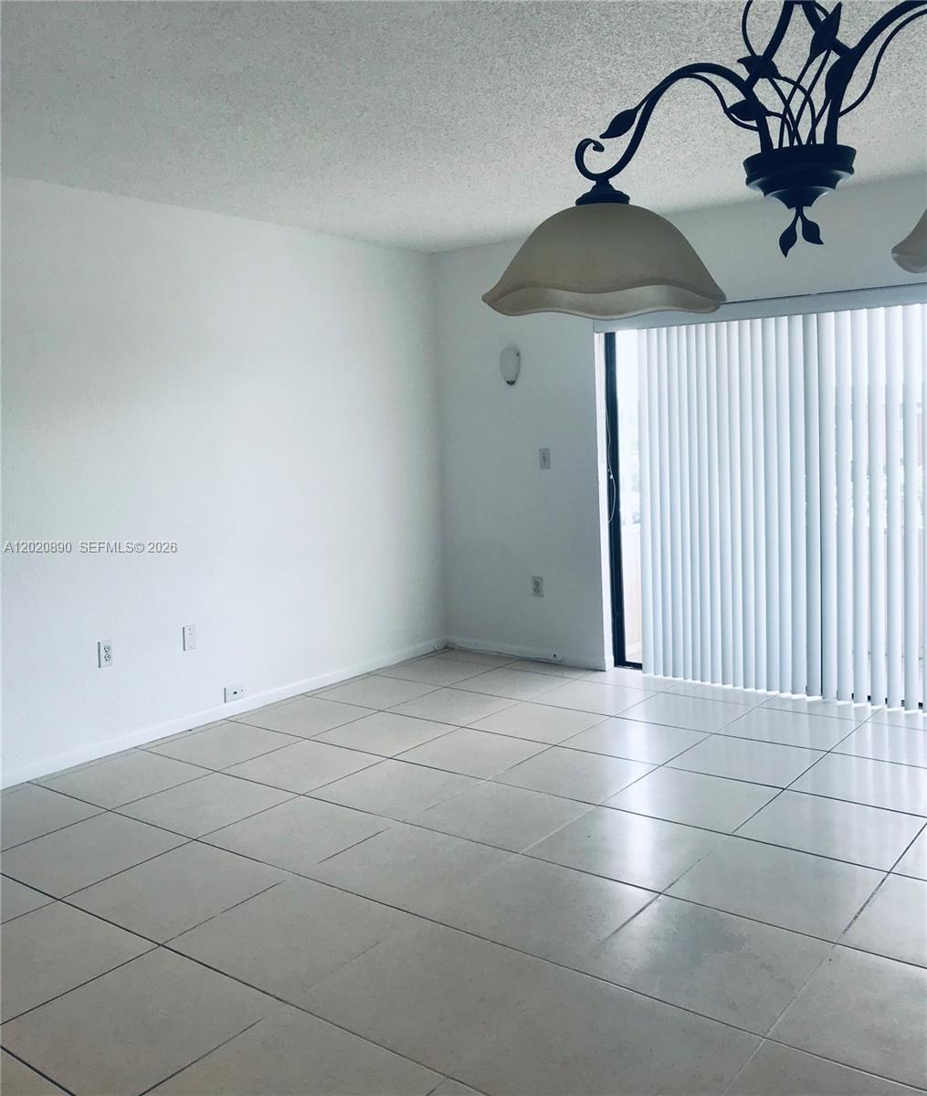 An empty room features white walls, light-colored tile flooring, a chandelier, and vertical blinds — 17600 NW 68th Ave # B3006, Hialeah FL 33015, HIALEAH, FL
