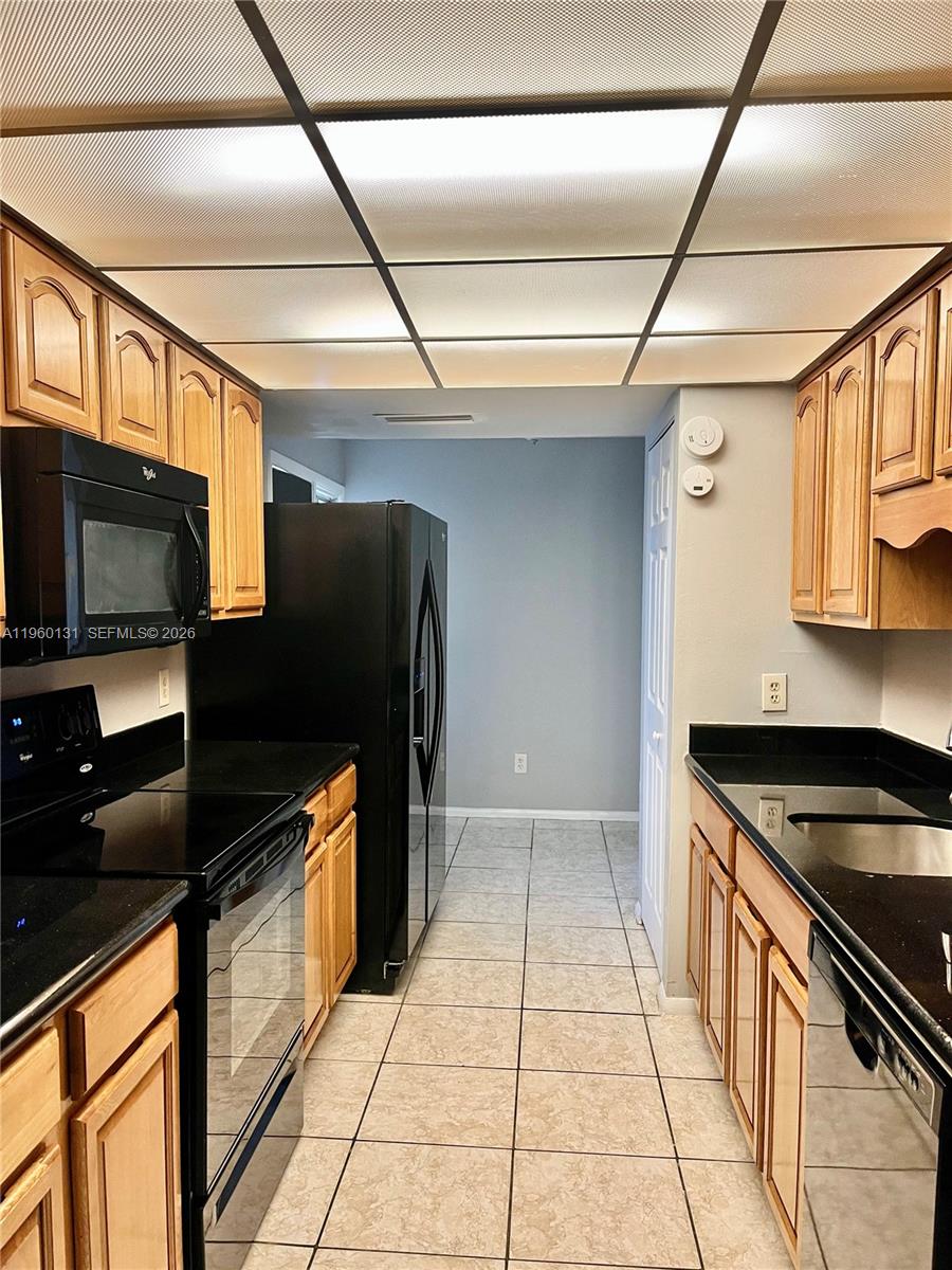 A galley kitchen features light wood cabinets, black appliances, black countertops, and a tiled floor — 4575 S Texas Ave # 307, Orlando FL 32839, ORLANDO, FL