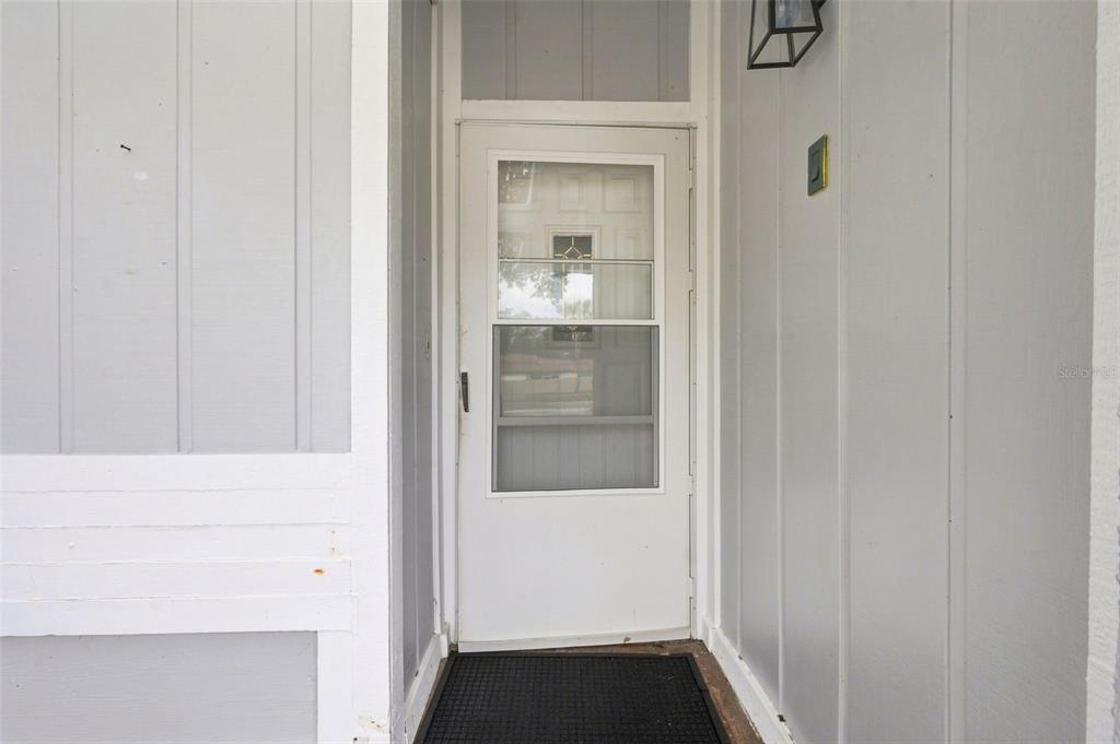 A white storm door with a glass panel is set within a light gray exterior wall — 1680 PINEHURST ROAD, DUNEDIN, FL