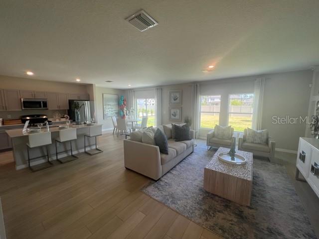 An open-concept living room and kitchen feature light wood flooring, neutral furniture, and large windows — 7568 HYDRANGEA LANE, WESLEY CHAPEL, FL