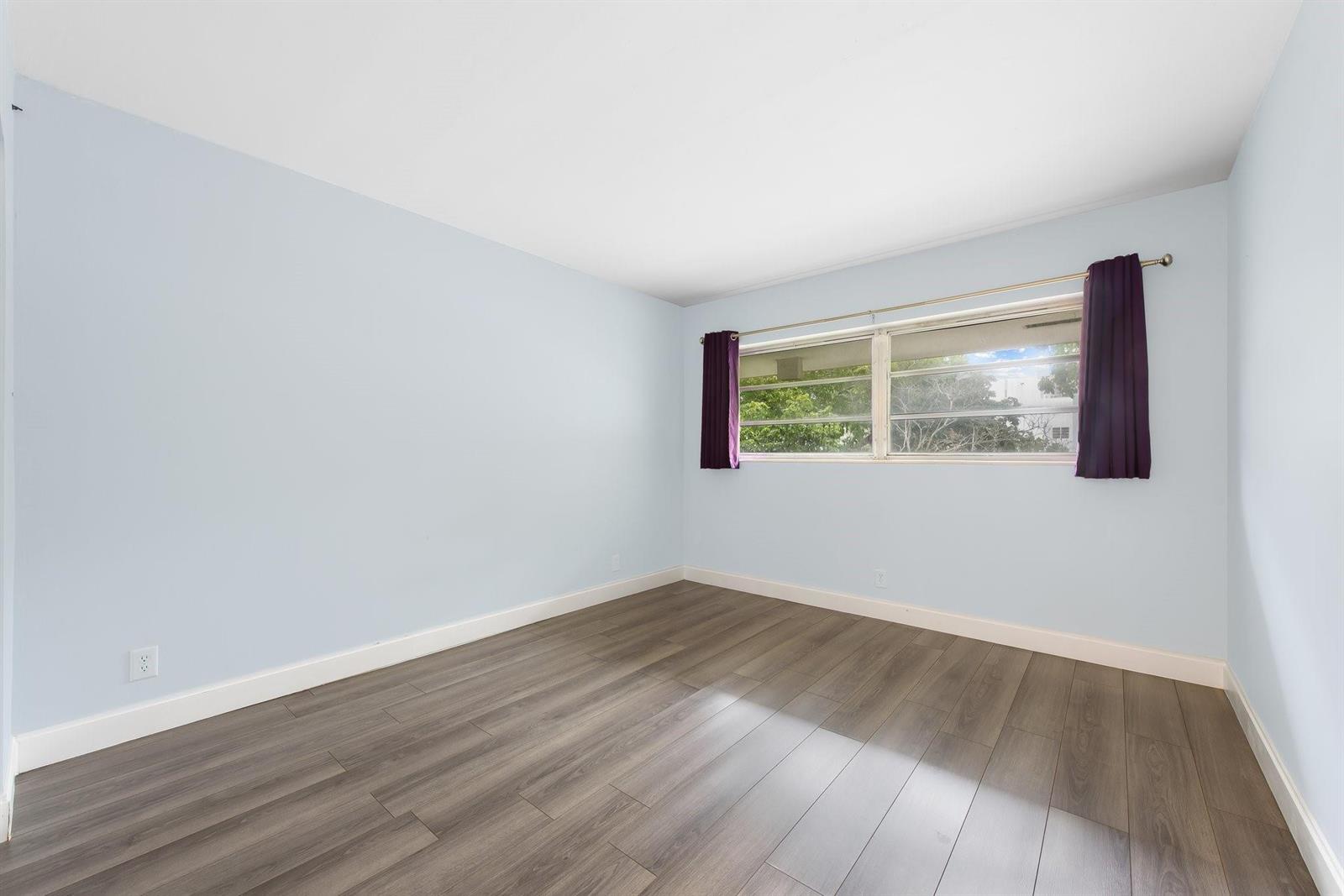 An empty bedroom features light blue walls, dark wood-look flooring, and a window with purple curtains — 1340 NE 28th Avenue # 232, Pompano Beach FL 33062, POMPANO BEACH, FL
