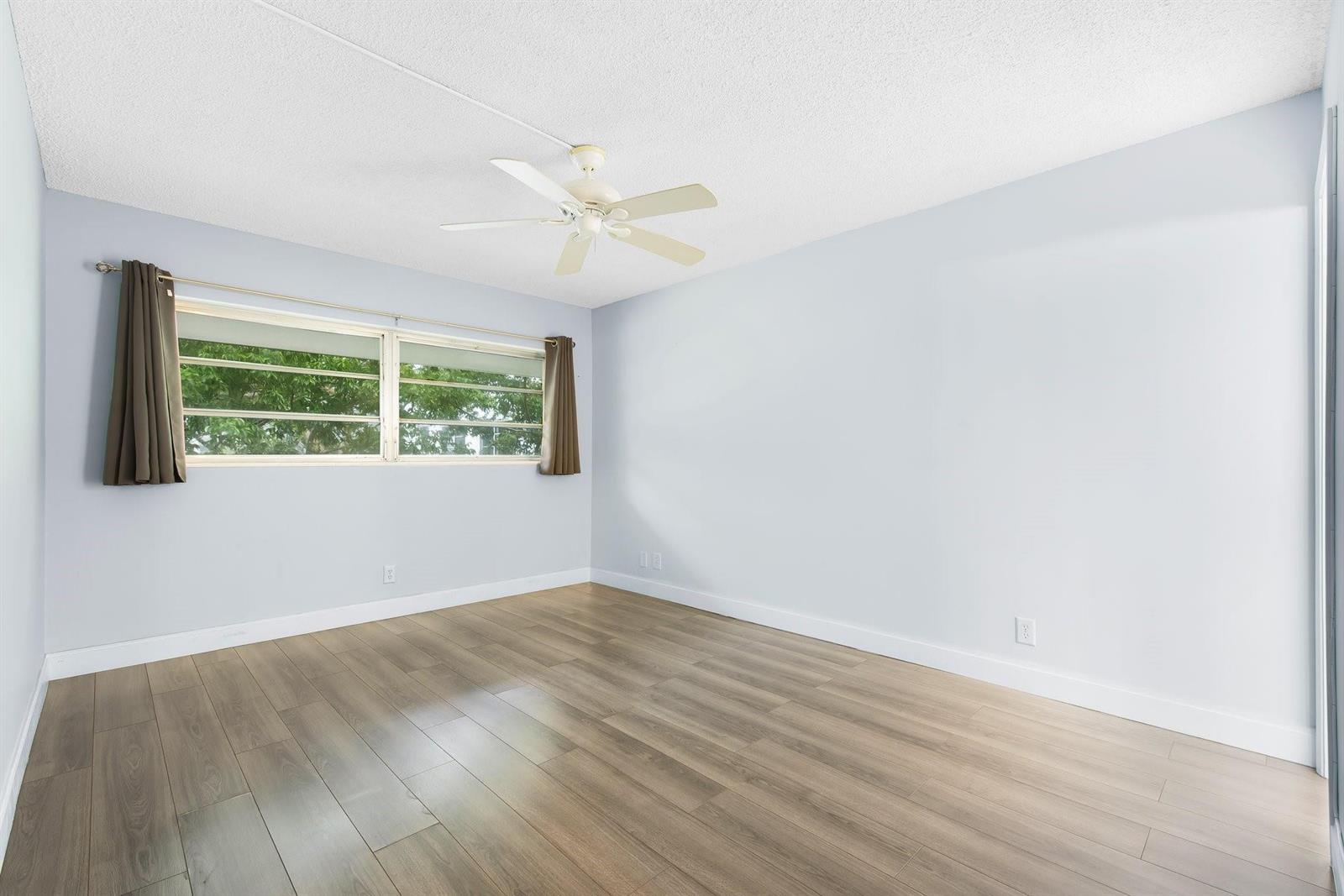 An empty bedroom features light blue walls, wood-look flooring, a ceiling fan, and a window — 1340 NE 28th Avenue # 232, Pompano Beach FL 33062, POMPANO BEACH, FL