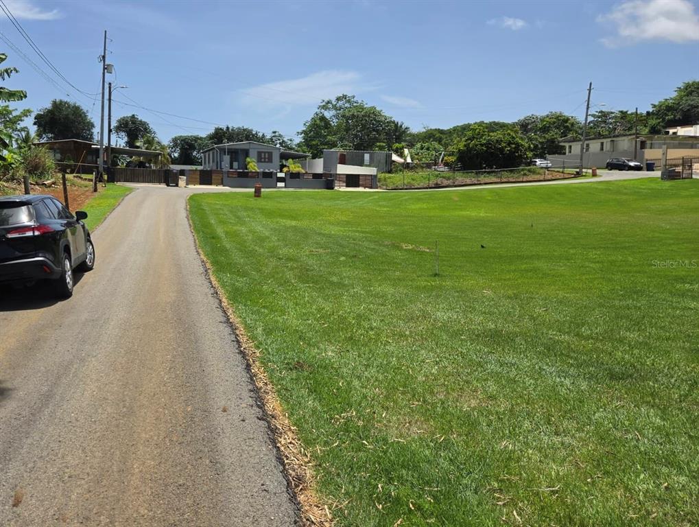 A paved road runs alongside a large, open grassy lot with houses in the background — Carr. 2, Km. 121.8 BO. CAIMITAL ALTO, AGUADILLA, PR