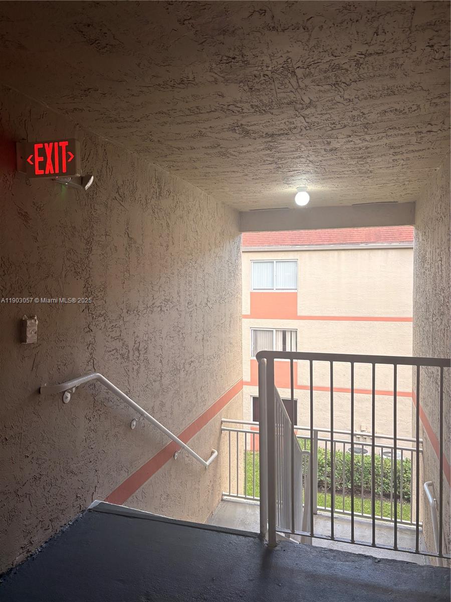 An outdoor stairwell features a red exit sign, white metal railings, and textured beige walls — 18850 NW 57th Ave # 306, Hialeah FL 33015, HIALEAH, FL