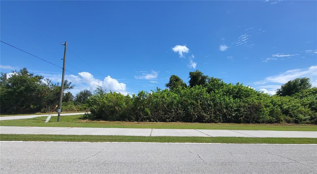 A vacant, overgrown lot sits behind a sidewalk and paved road under a blue sky — 10044 CALUMET BOULEVARD, PORT CHARLOTTE, FL