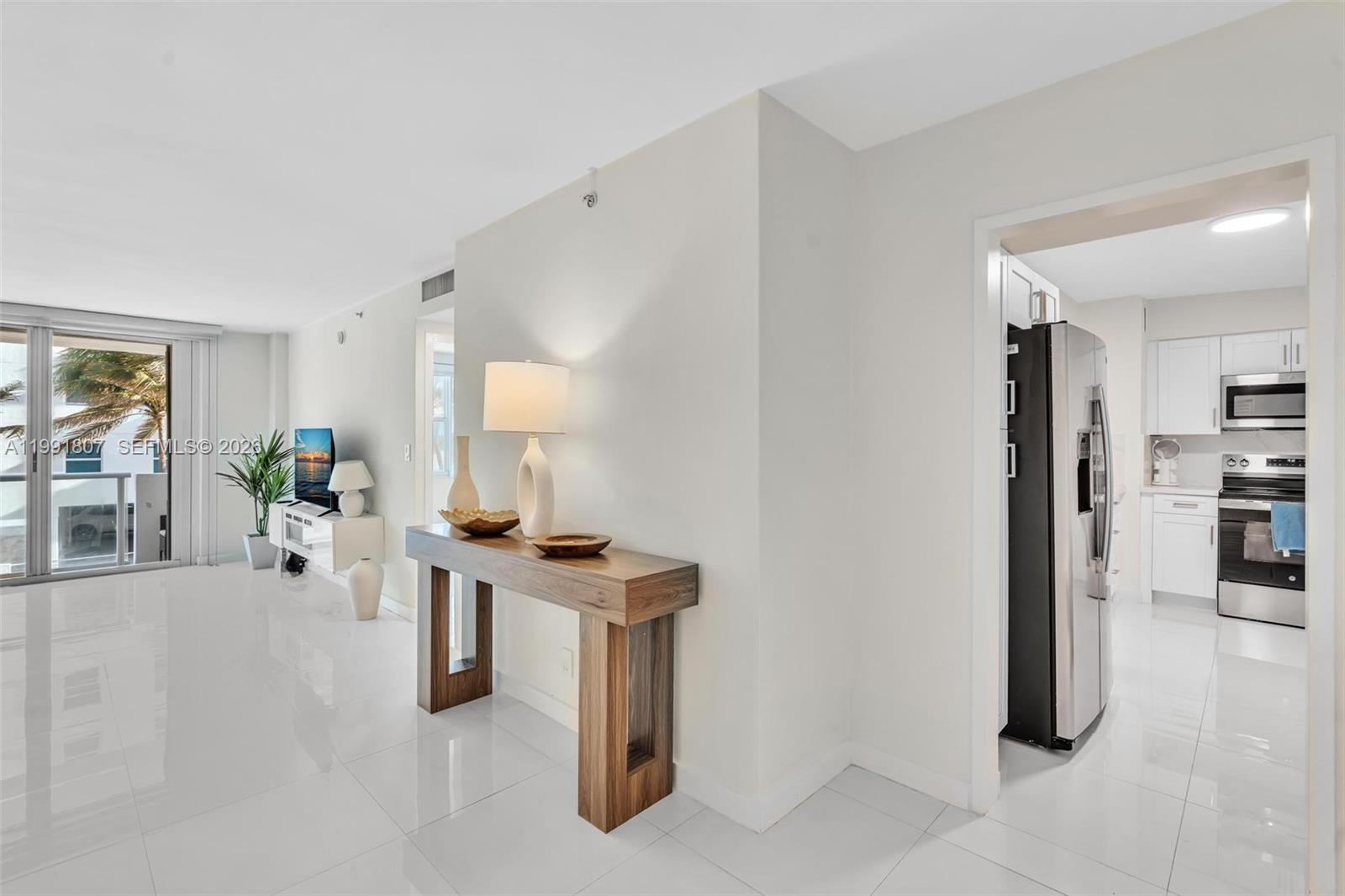 A bright living area with white tile floors leads to a kitchen and balcony — 9195 Collins Ave # 303, Surfside FL 33154, SURFSIDE, FL