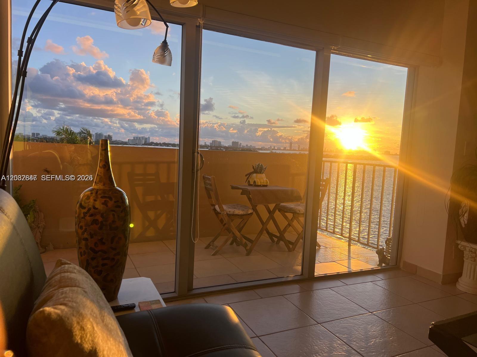A living room with tile floors overlooks a balcony and water view at sunset — 880 NE 69th St # 6R, Miami FL 33138, MIAMI, FL