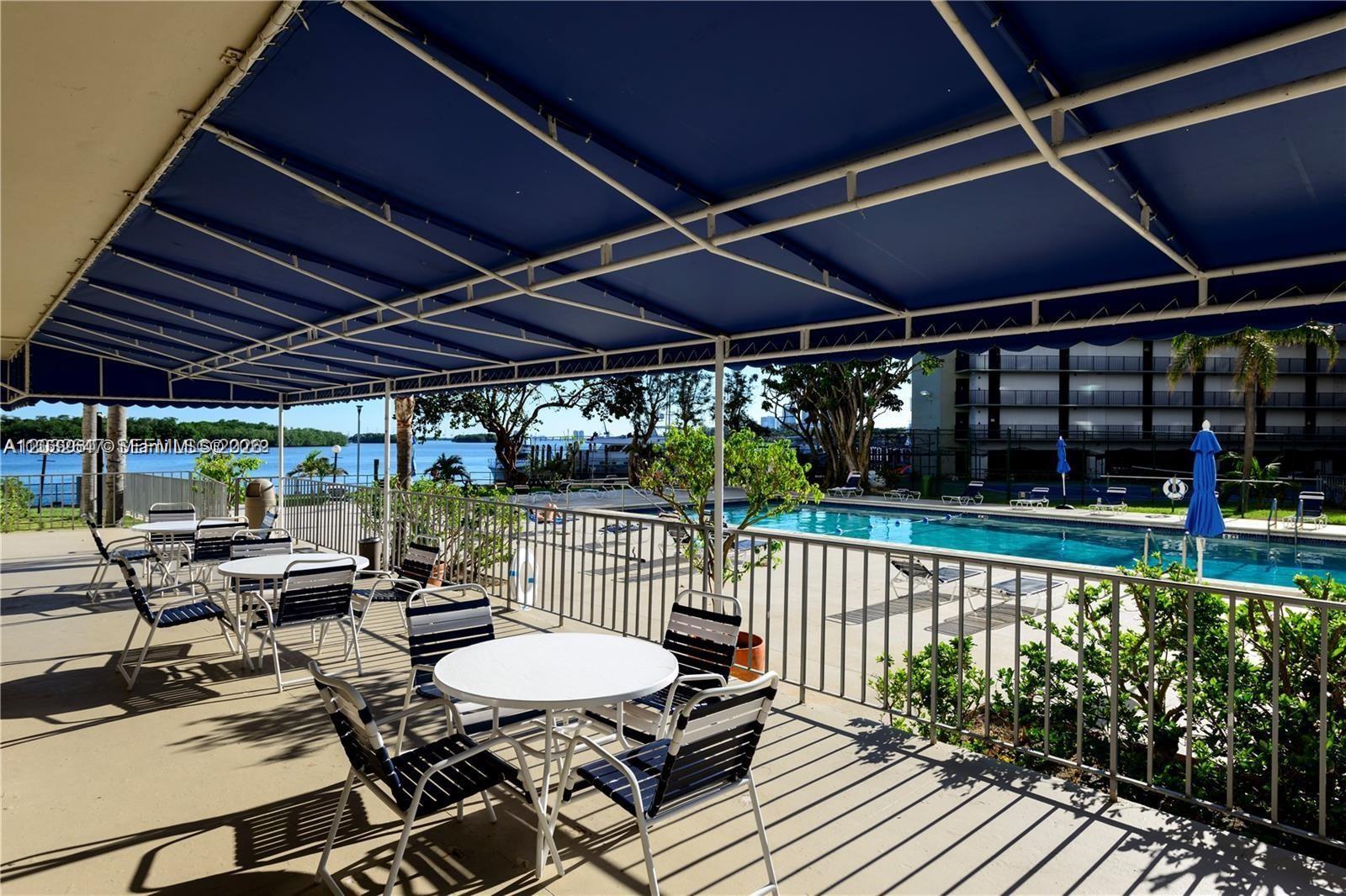 A covered patio with tables and chairs overlooks a swimming pool and a waterfront view — 880 NE 69th St # 6R, Miami FL 33138, MIAMI, FL