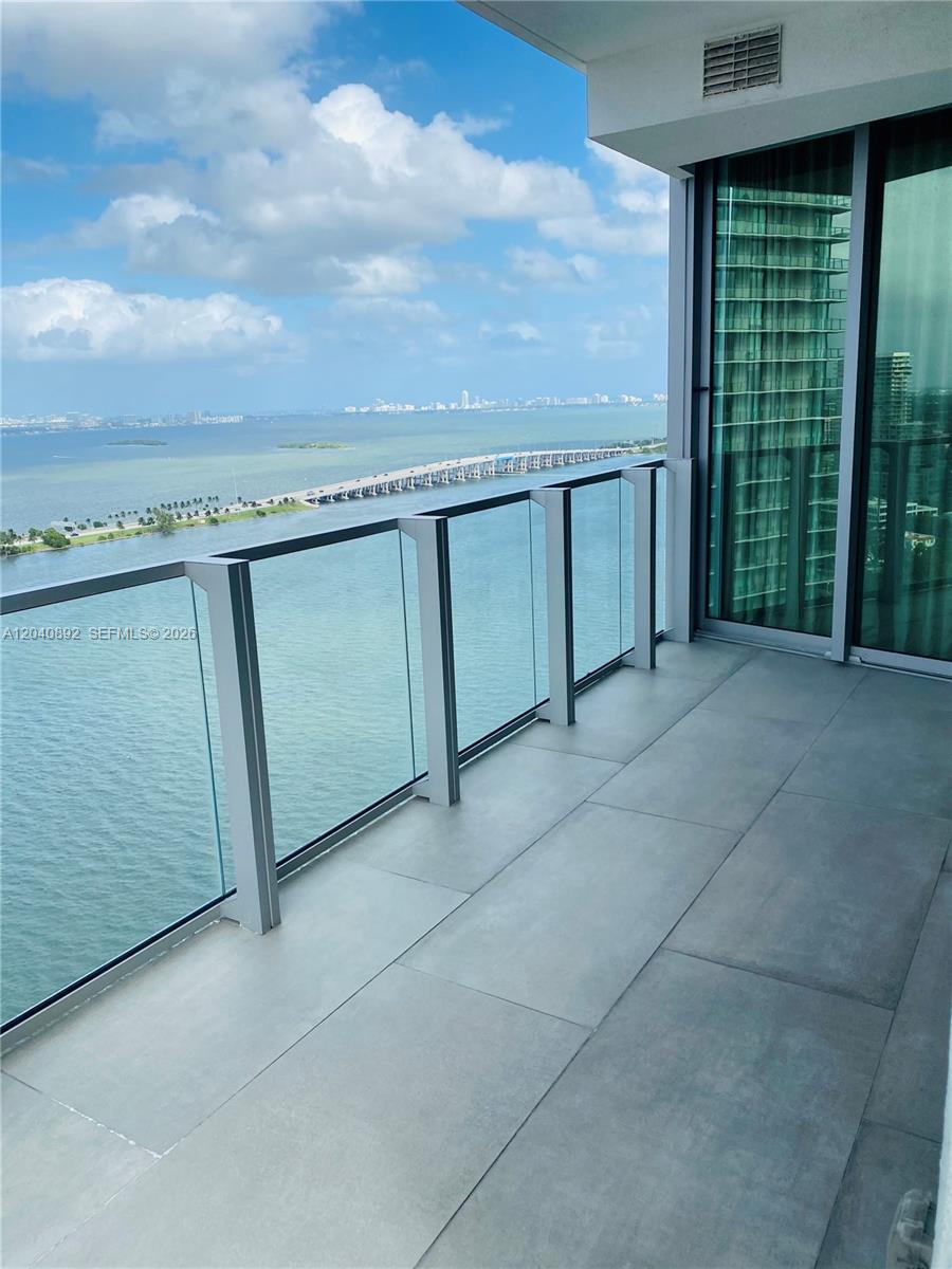 A balcony with gray tile flooring and a glass railing overlooks a bay and bridge — MIAMI, FL