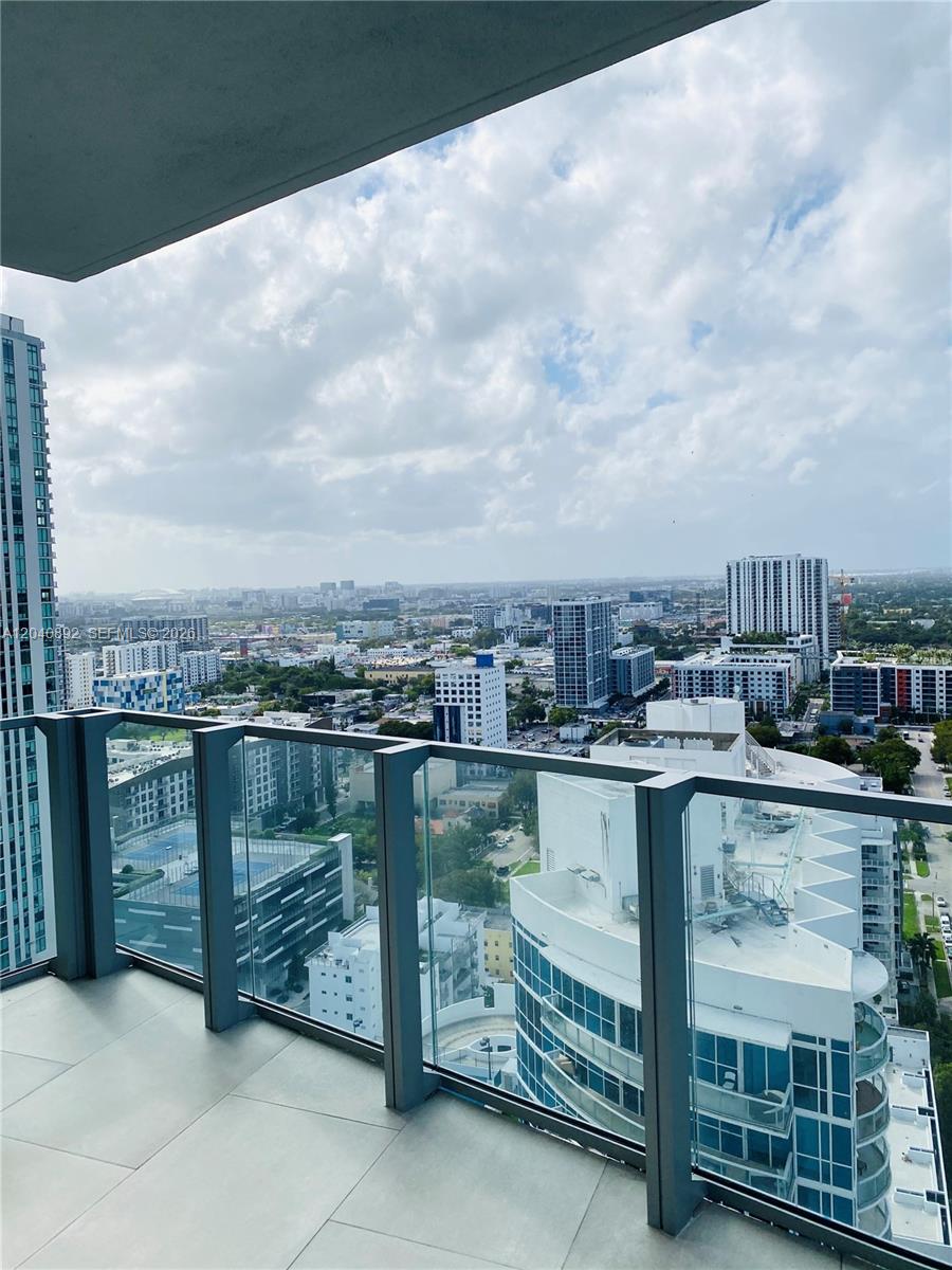 A high-rise balcony with glass railings overlooks a dense urban cityscape under a cloudy sky — MIAMI, FL