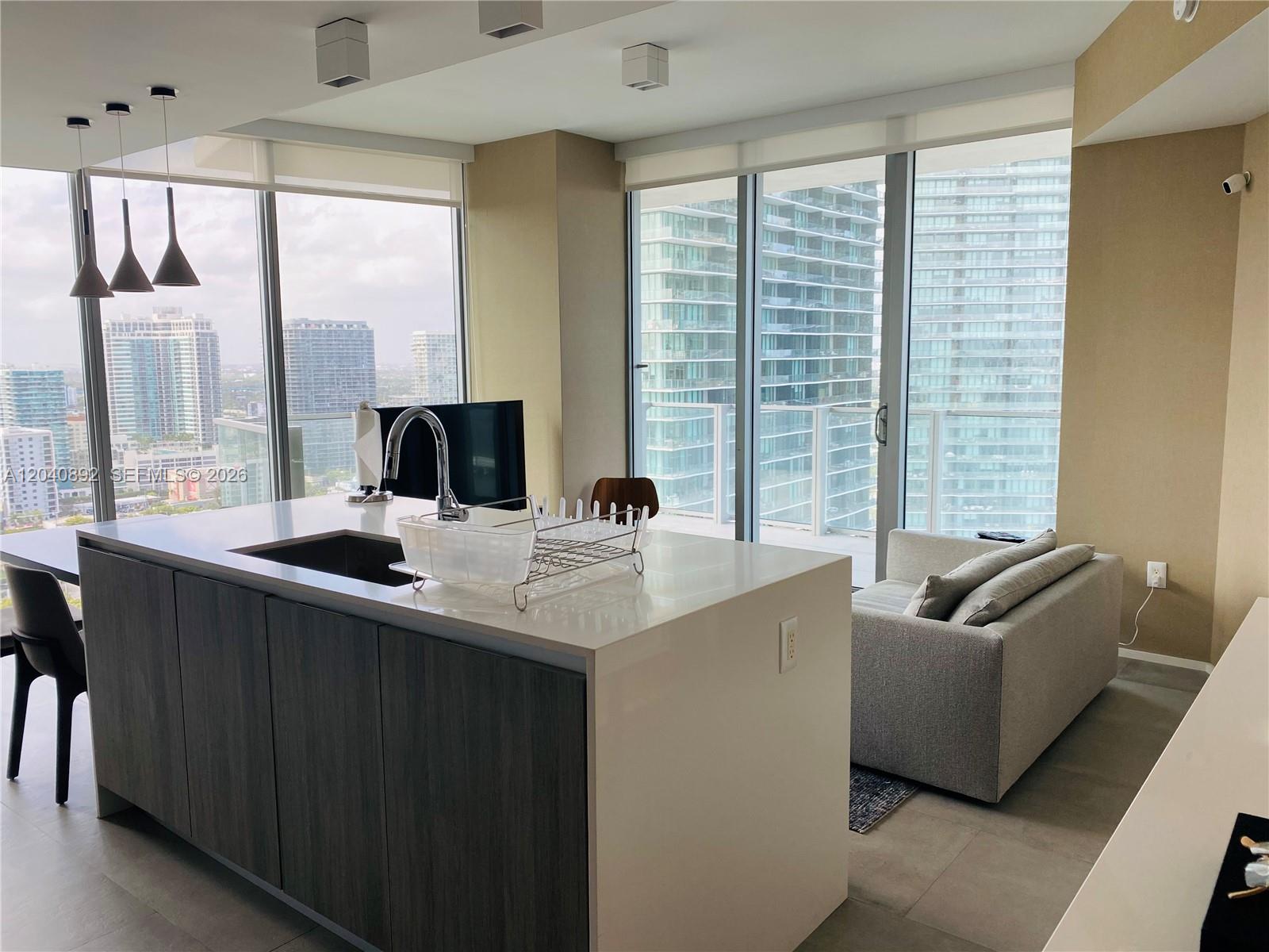 A modern kitchen island with a sink sits before a living area and floor-to-ceiling windows — MIAMI, FL